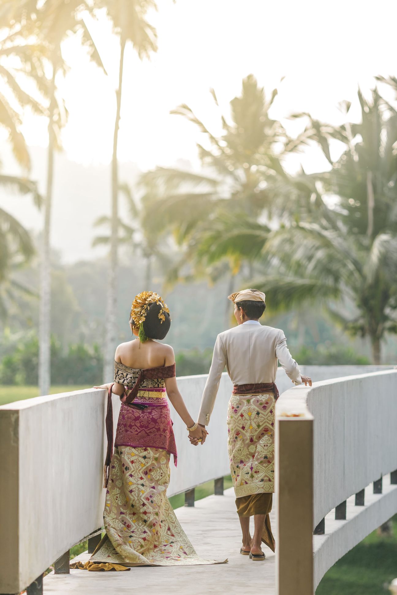 A couple in traditional Balinese wedding attire walk hand in hand along a sunlit bridge