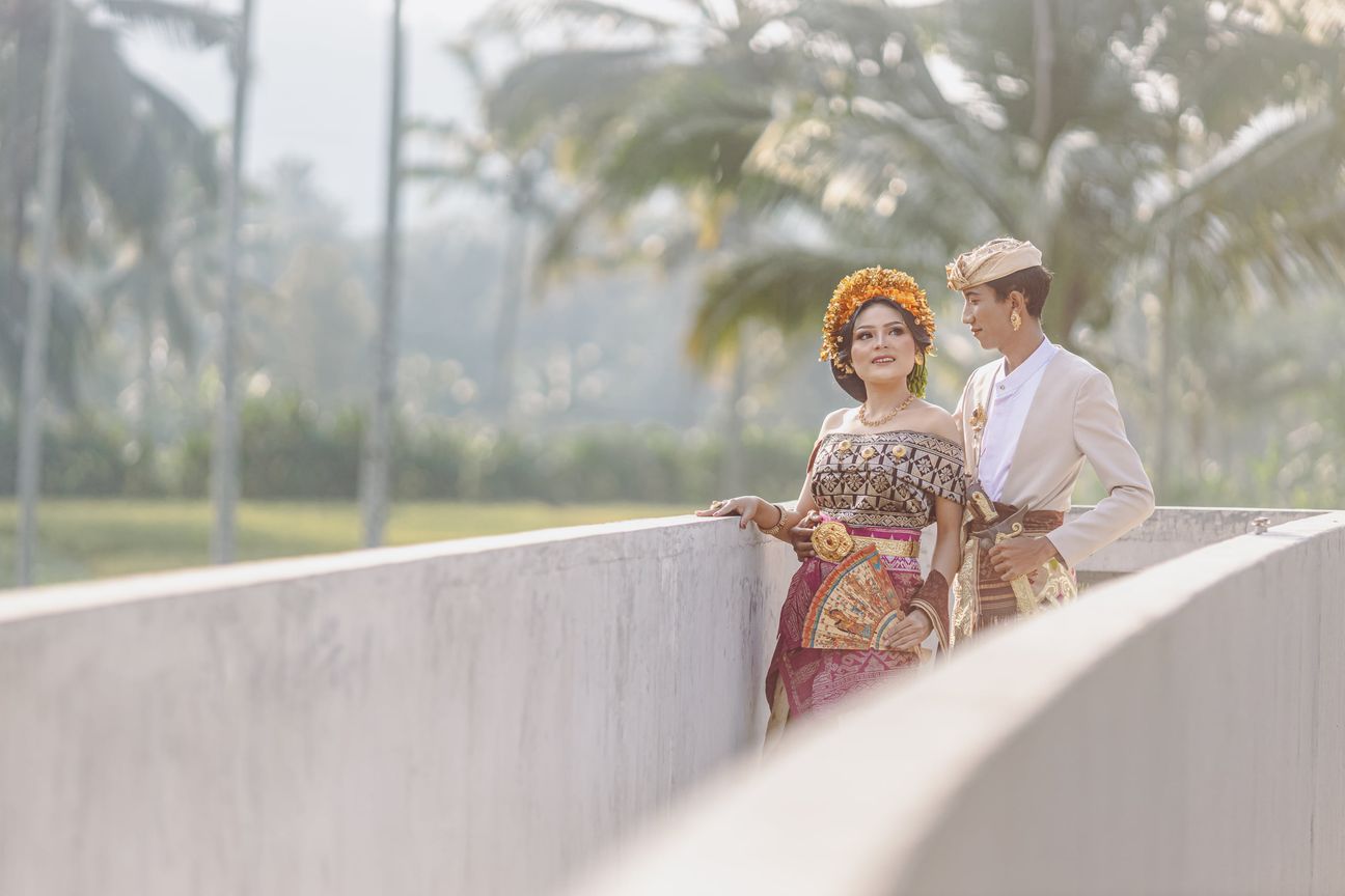 A couple in traditional Balinese wedding attire walk hand in hand along a sunlit bridge