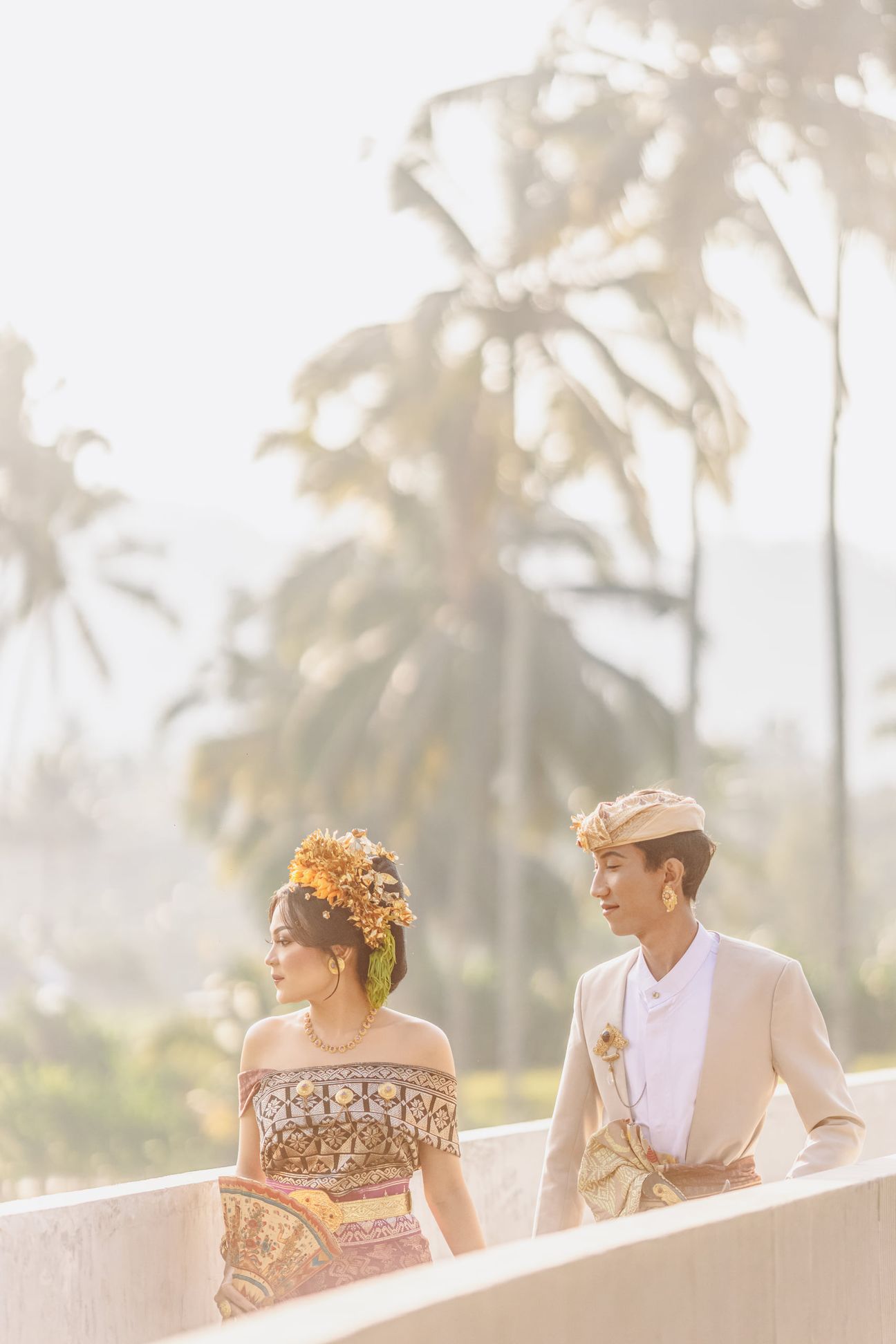 A couple in traditional Balinese wedding attire walk hand in hand along a sunlit bridge
