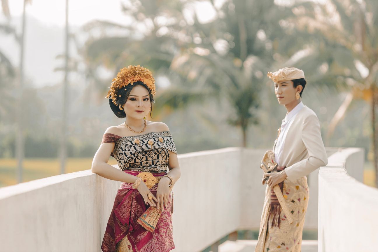 A couple in traditional Balinese wedding attire walk hand in hand along a sunlit bridge