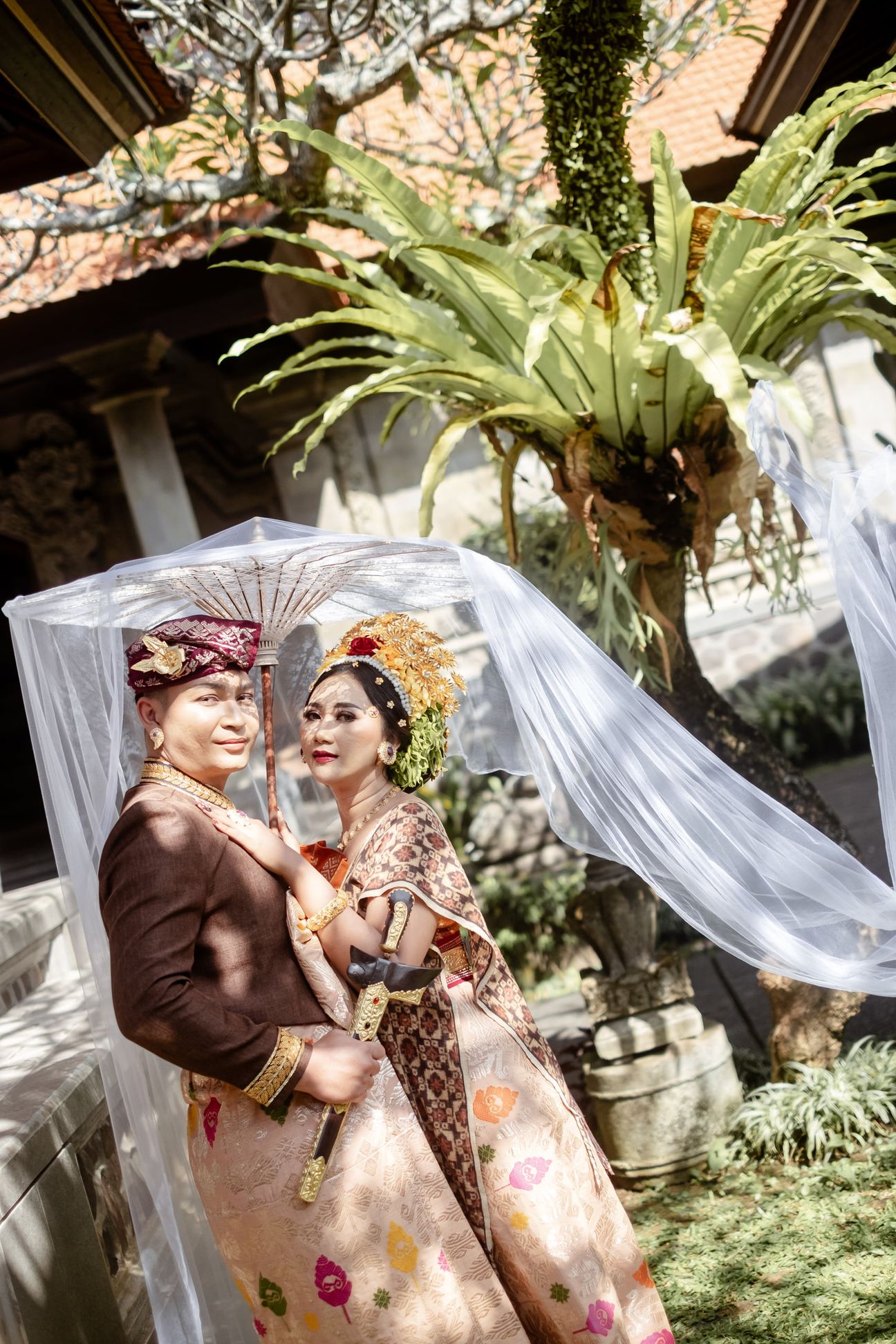 A couple in traditional Balinese wedding dress pose beneath a white ceremonial parasol