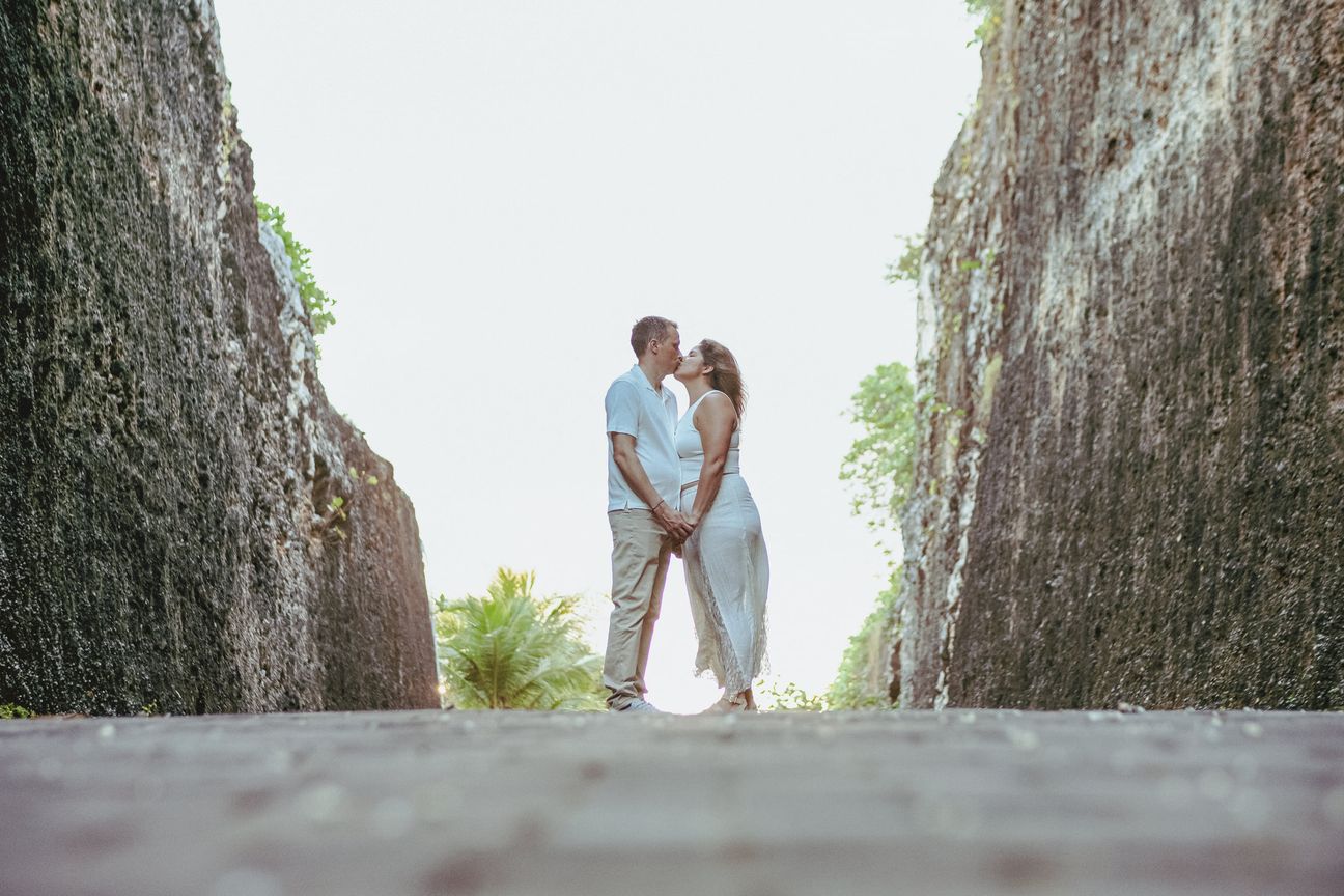A couple walk between tall stone cliffs in bright coastal light