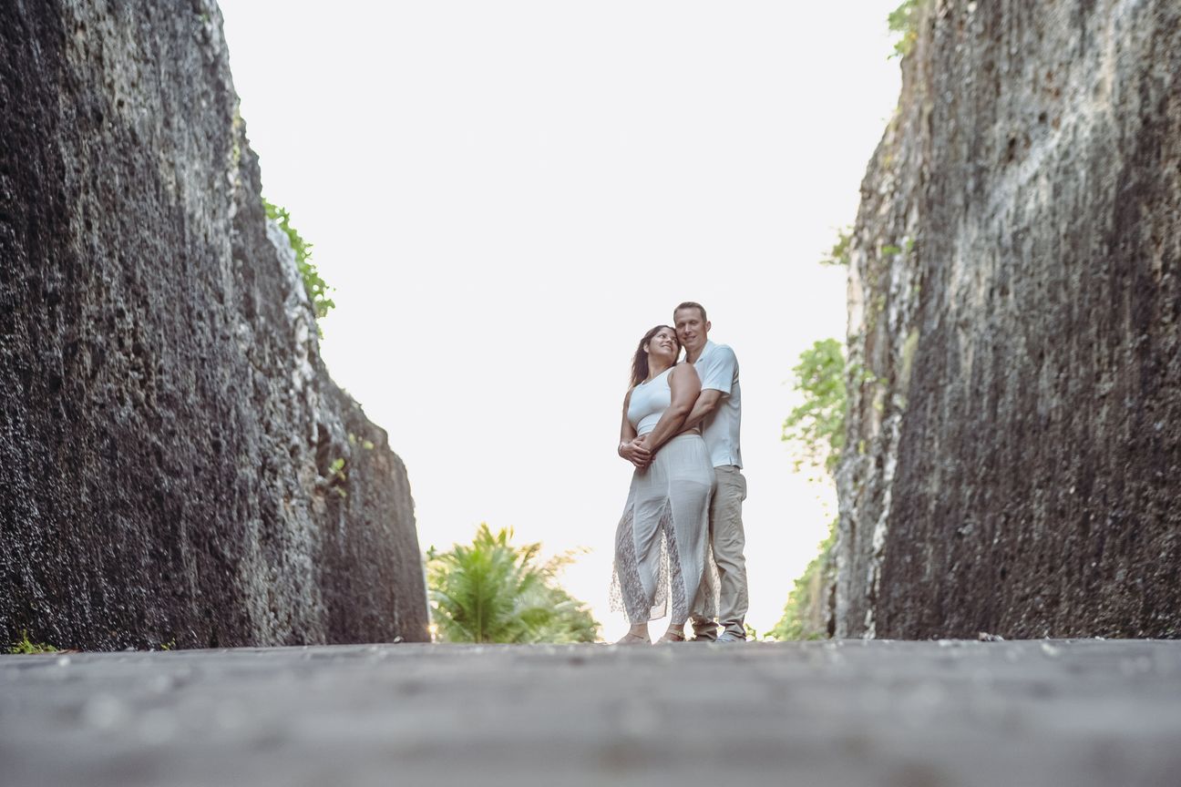 A couple walk between tall stone cliffs in bright coastal light