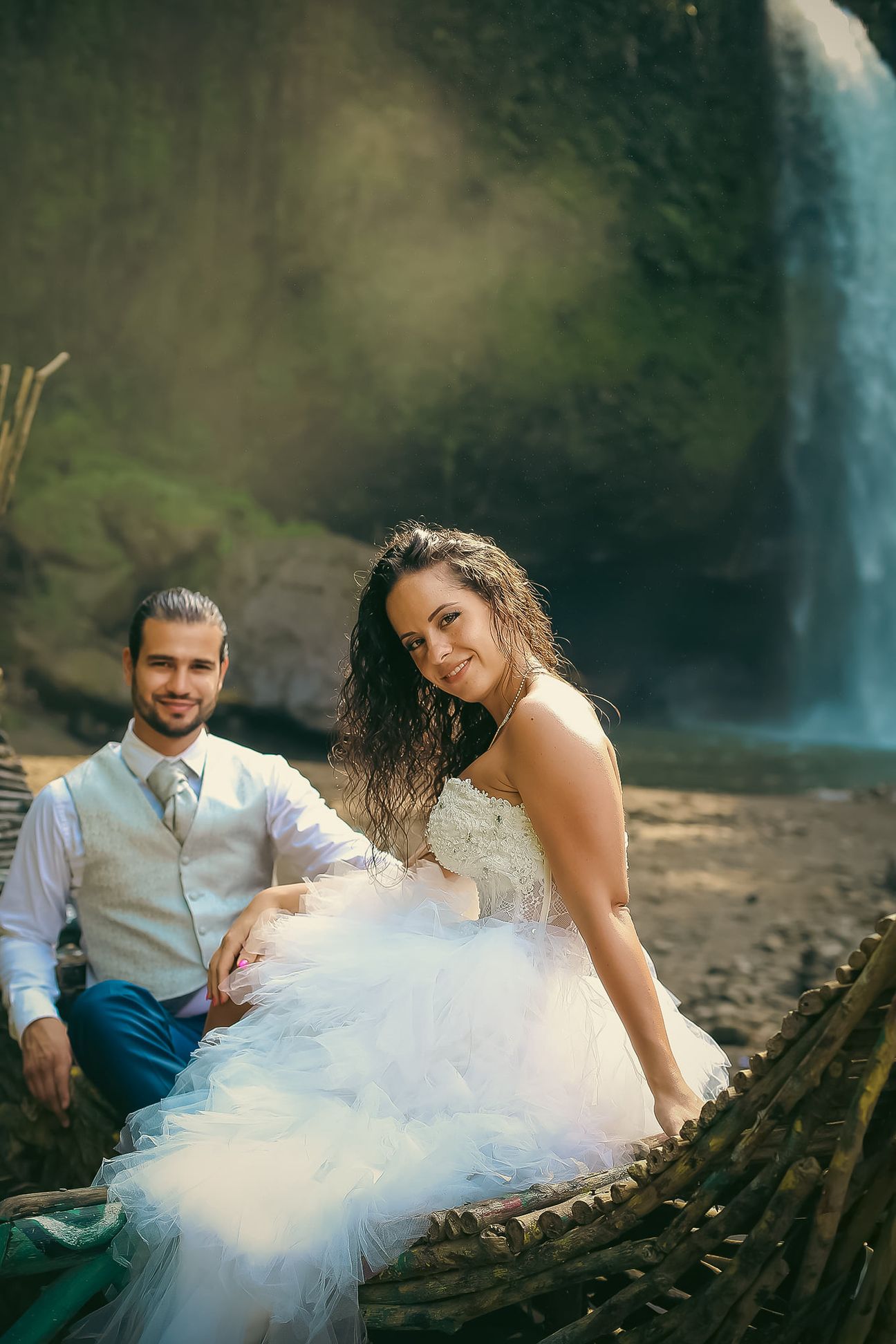 A newlywed couple sit together beside a forest waterfall