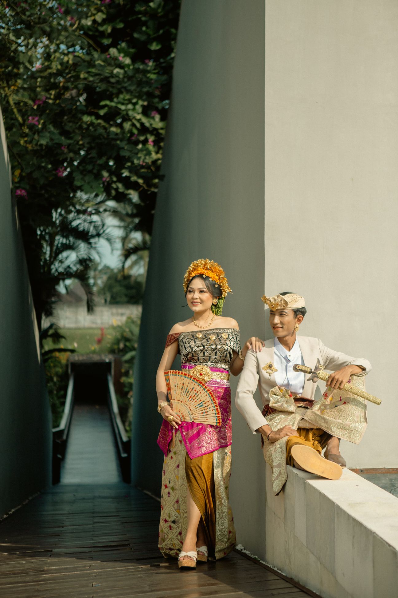 A couple in Balinese attire pause together beside a peaceful garden pavilion