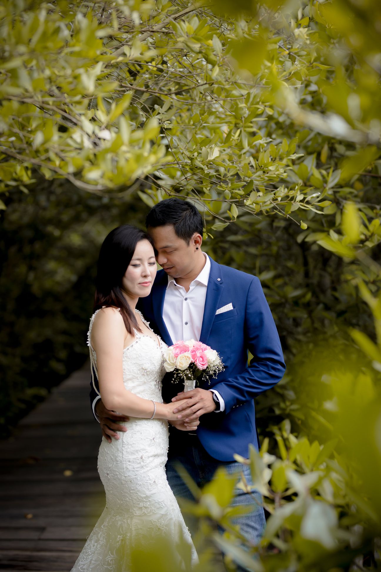 A bride and groom embrace along a wooden boardwalk through leafy mangroves