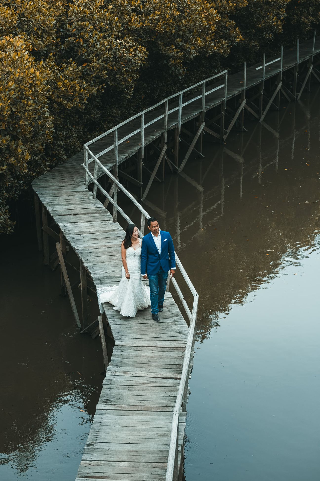 A bride and groom embrace along a wooden boardwalk through leafy mangroves