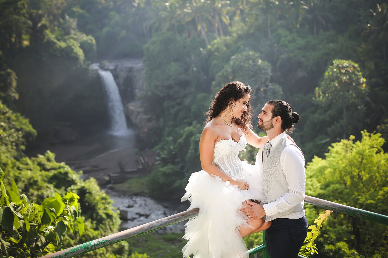 A newlywed couple pose together beneath a tall forest waterfall
