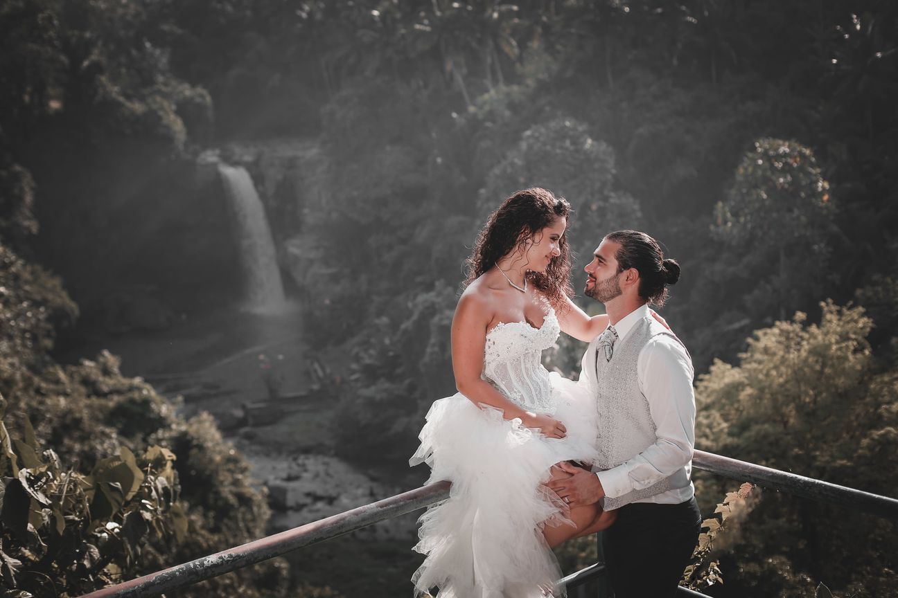 A newlywed couple pose together beneath a tall forest waterfall
