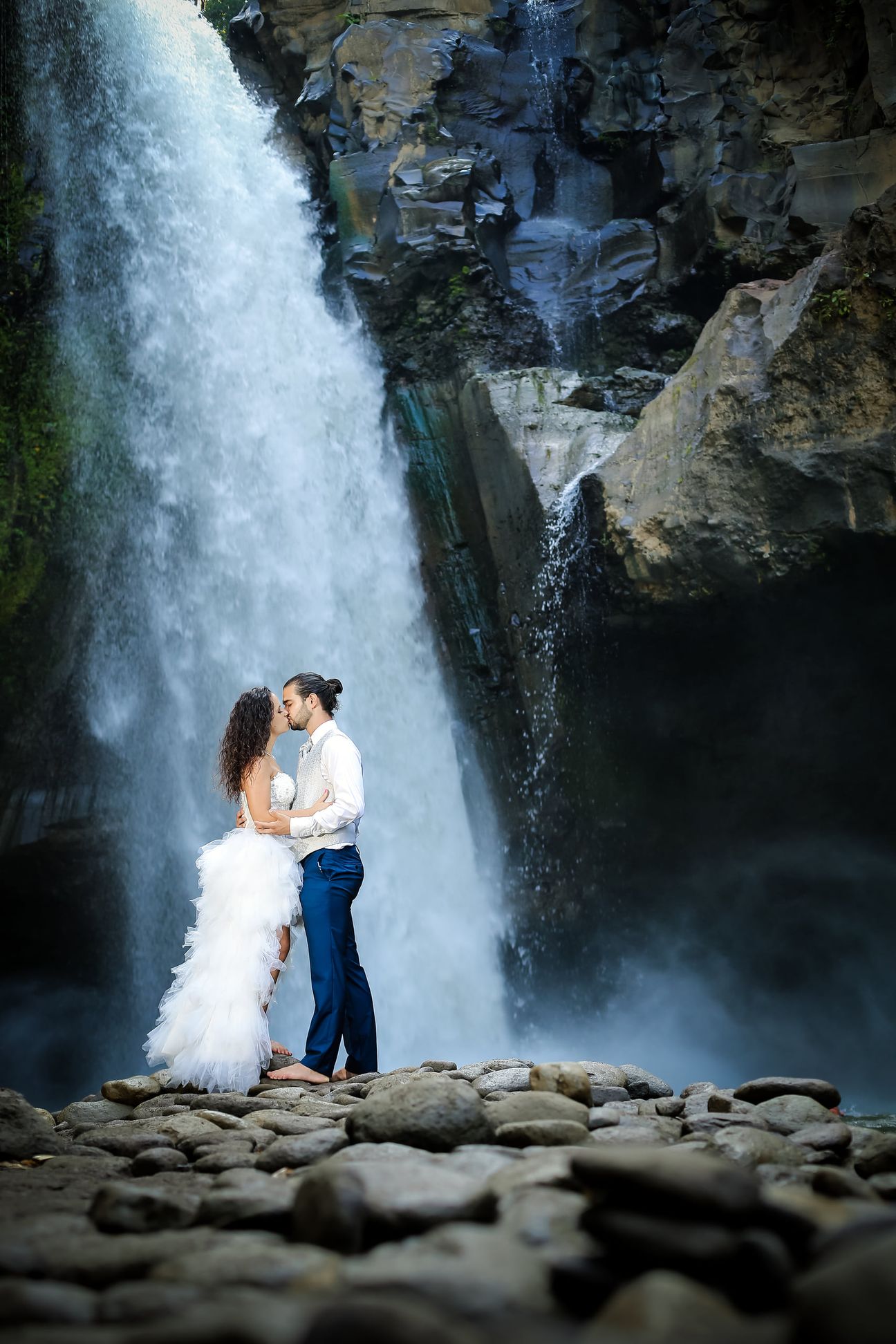 A newlywed couple pose together beneath a tall forest waterfall