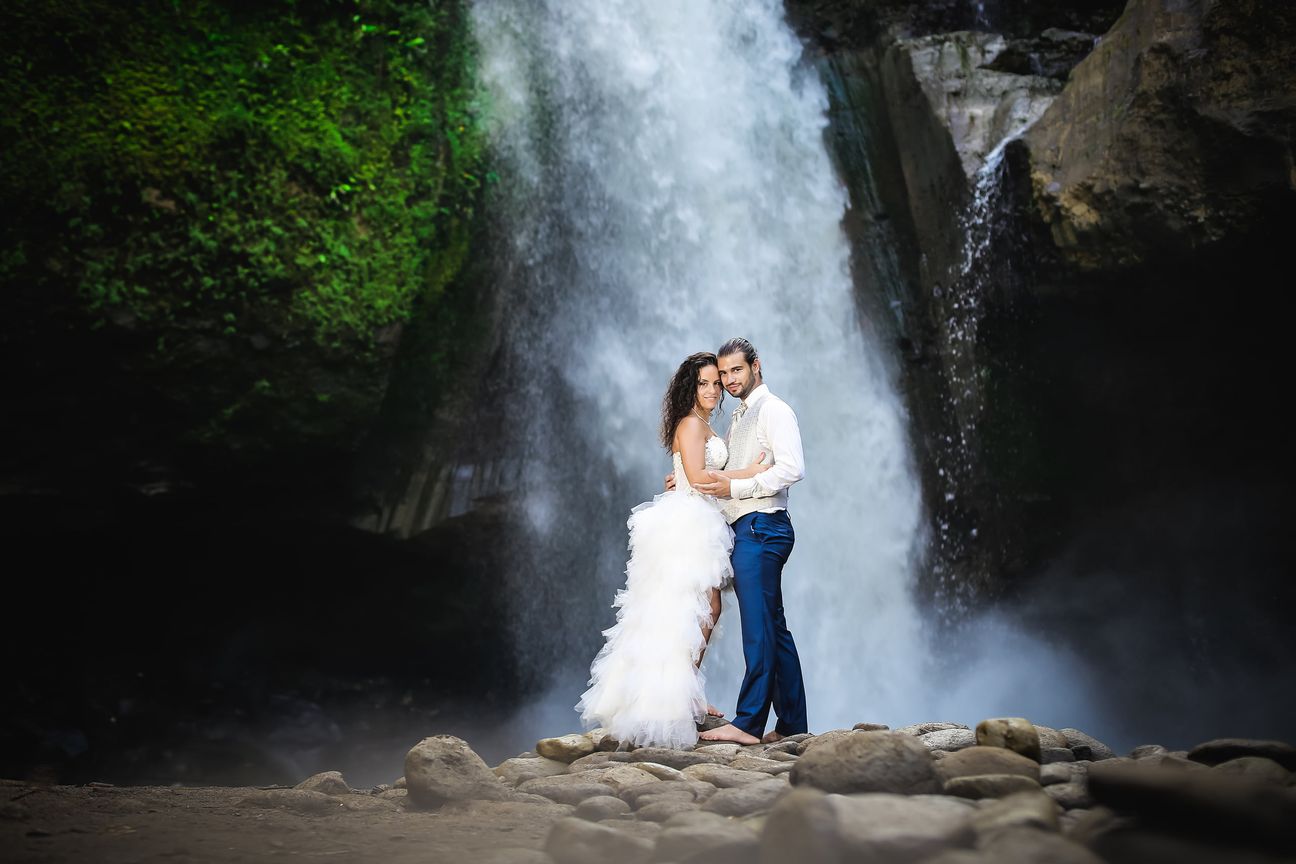 A newlywed couple pose together beneath a tall forest waterfall