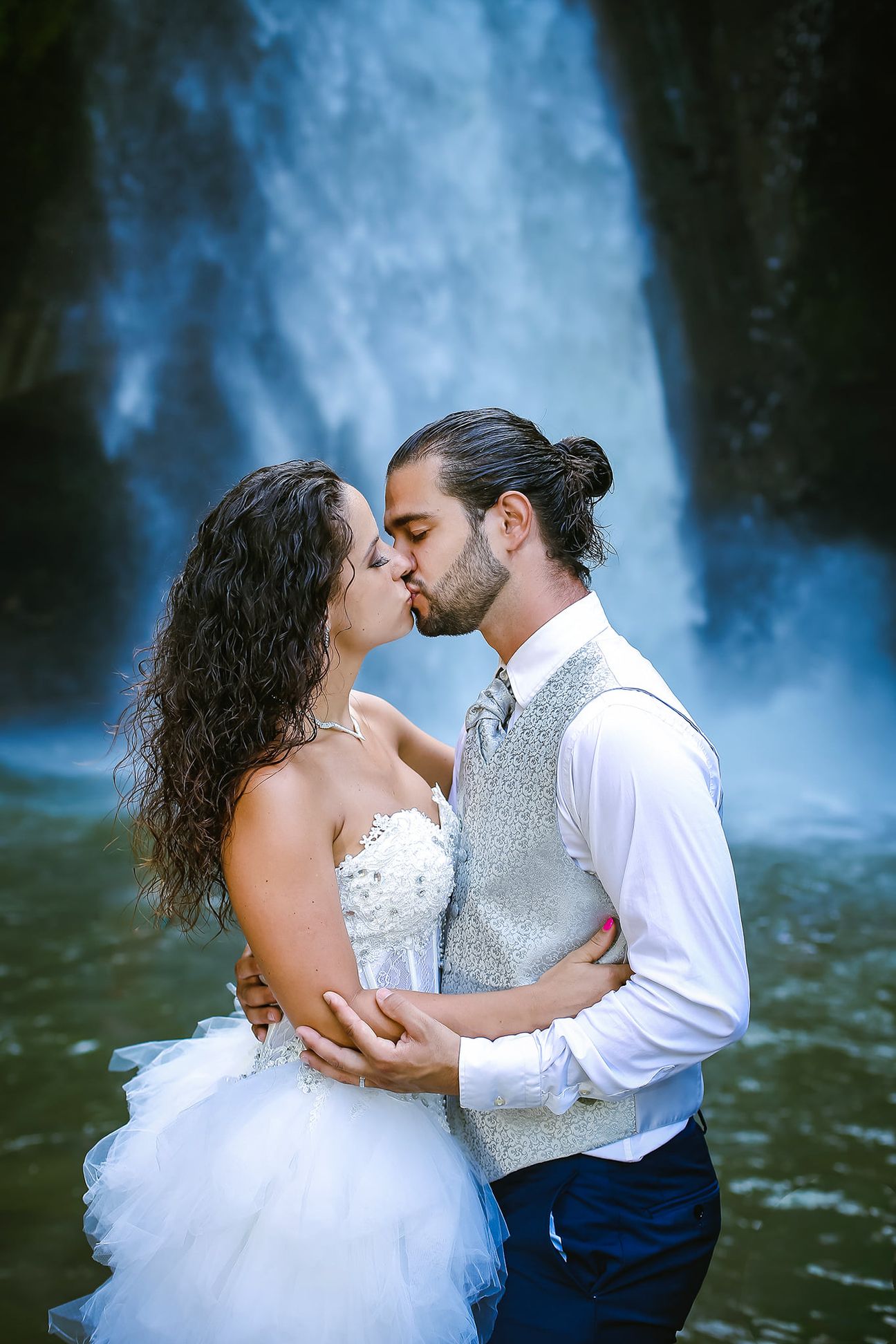 A newlywed couple pose together beneath a tall forest waterfall