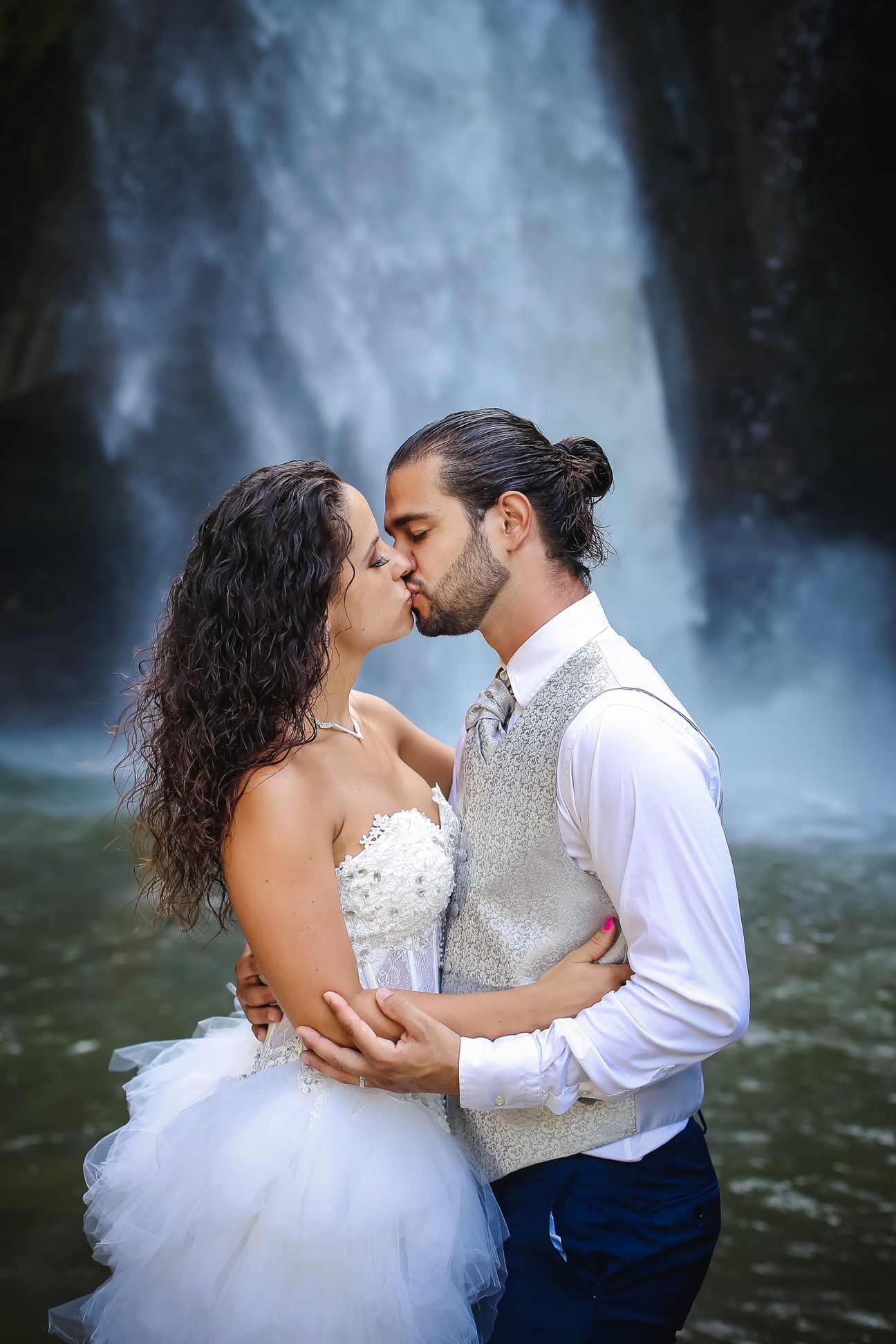 A newlywed couple pose together beneath a tall forest waterfall