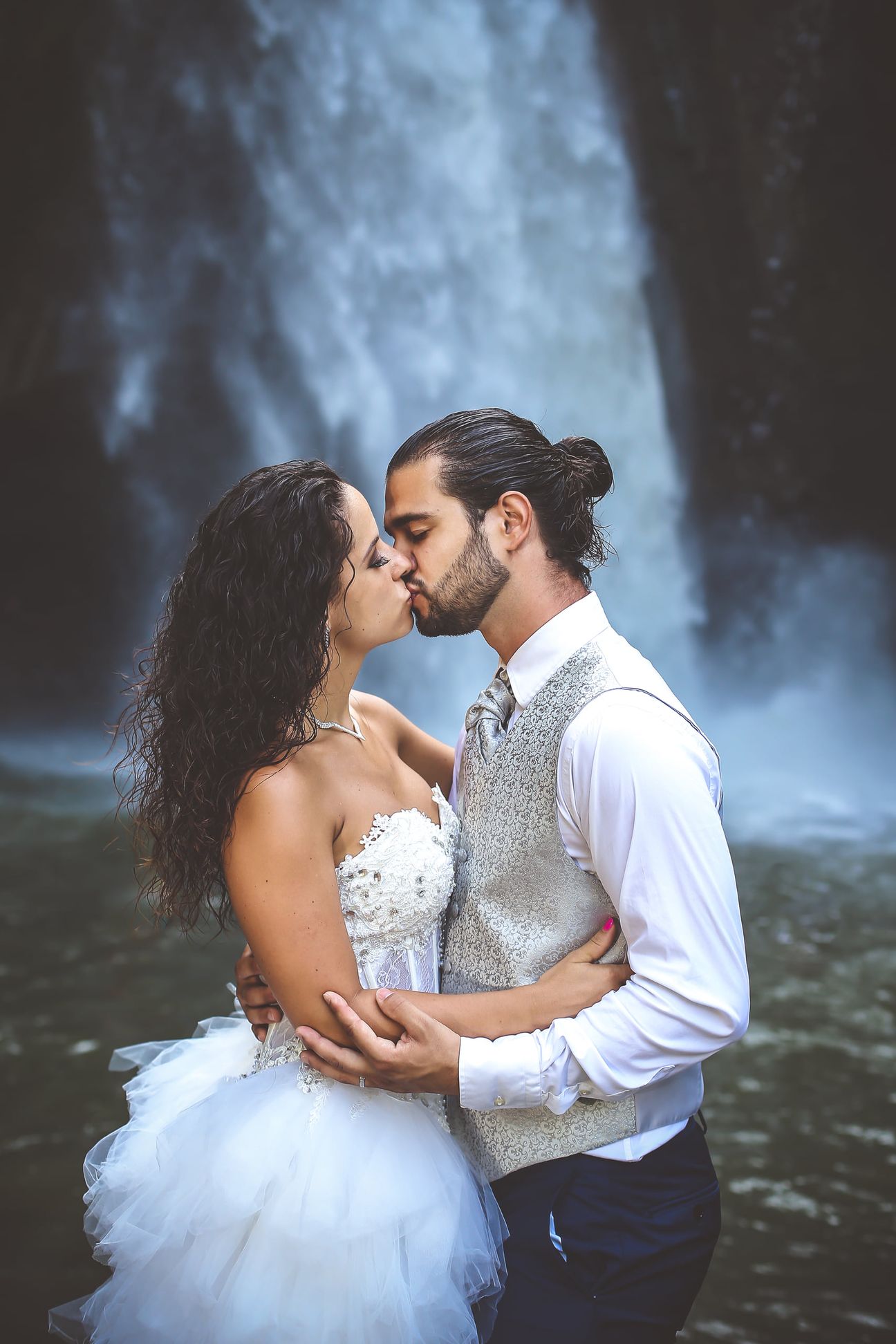 A newlywed couple pose together beneath a tall forest waterfall