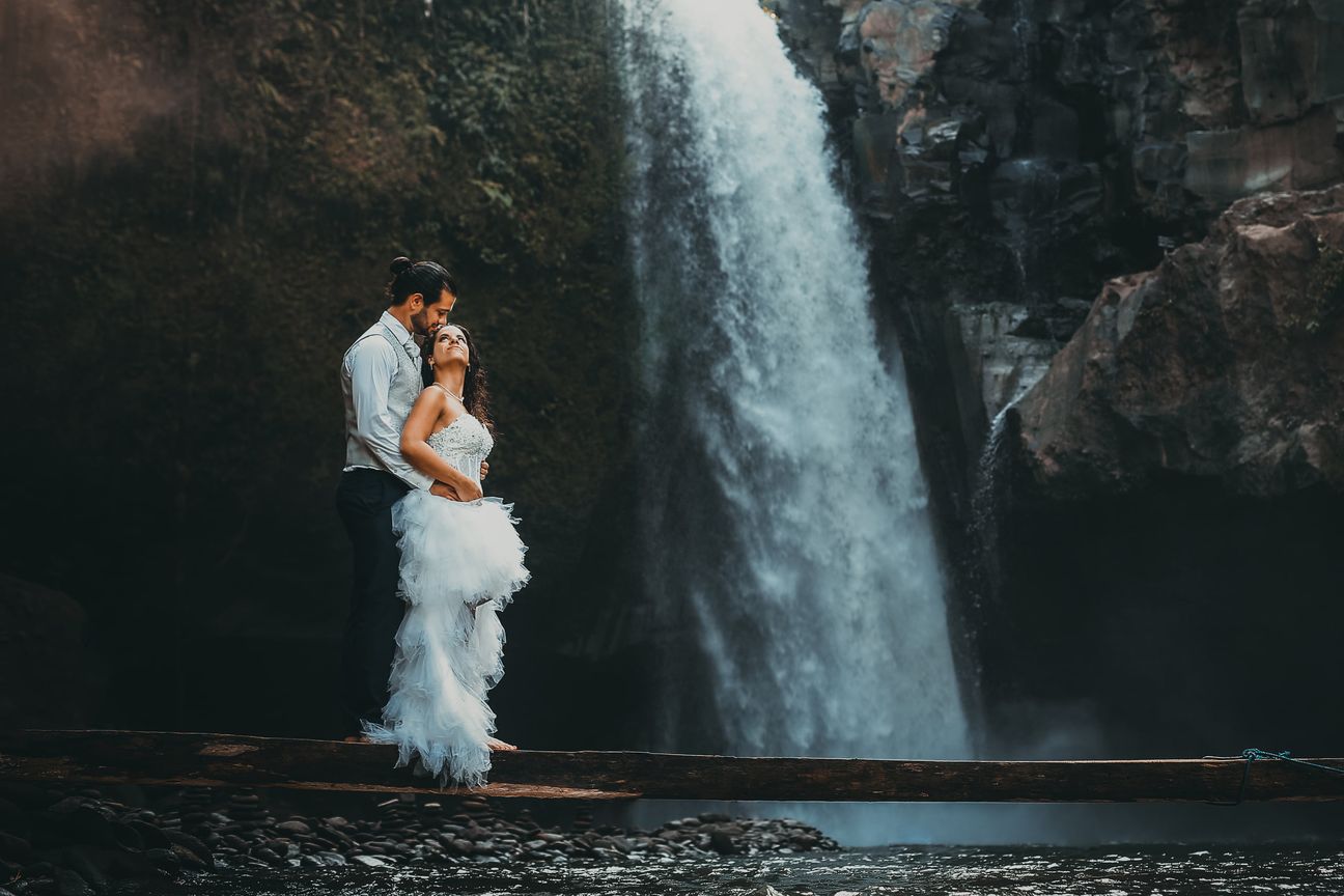 A newlywed couple pose together beneath a tall forest waterfall