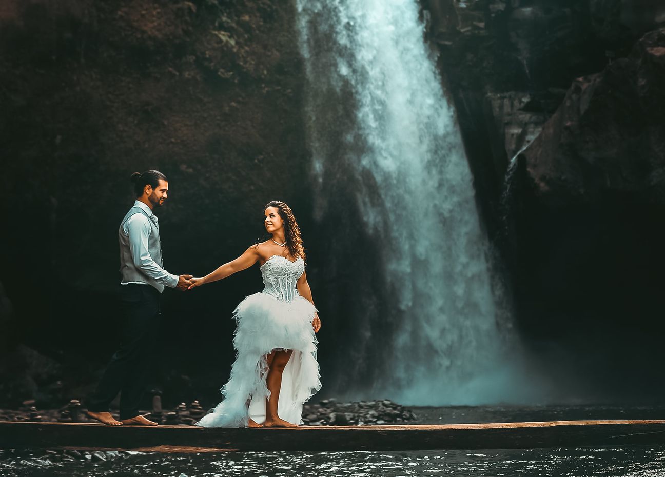 A newlywed couple pose together beneath a tall forest waterfall
