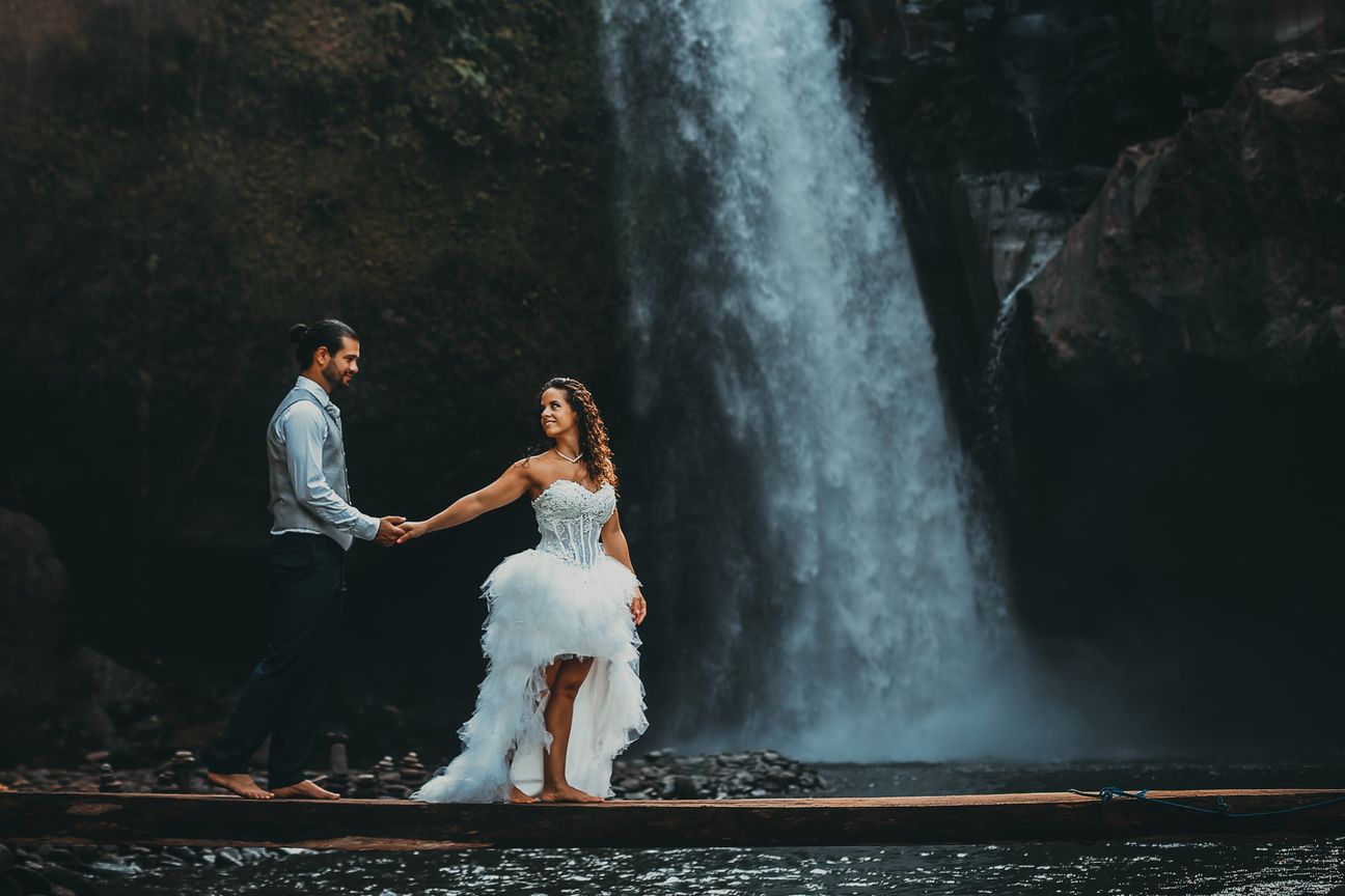 A newlywed couple pose together beneath a tall forest waterfall