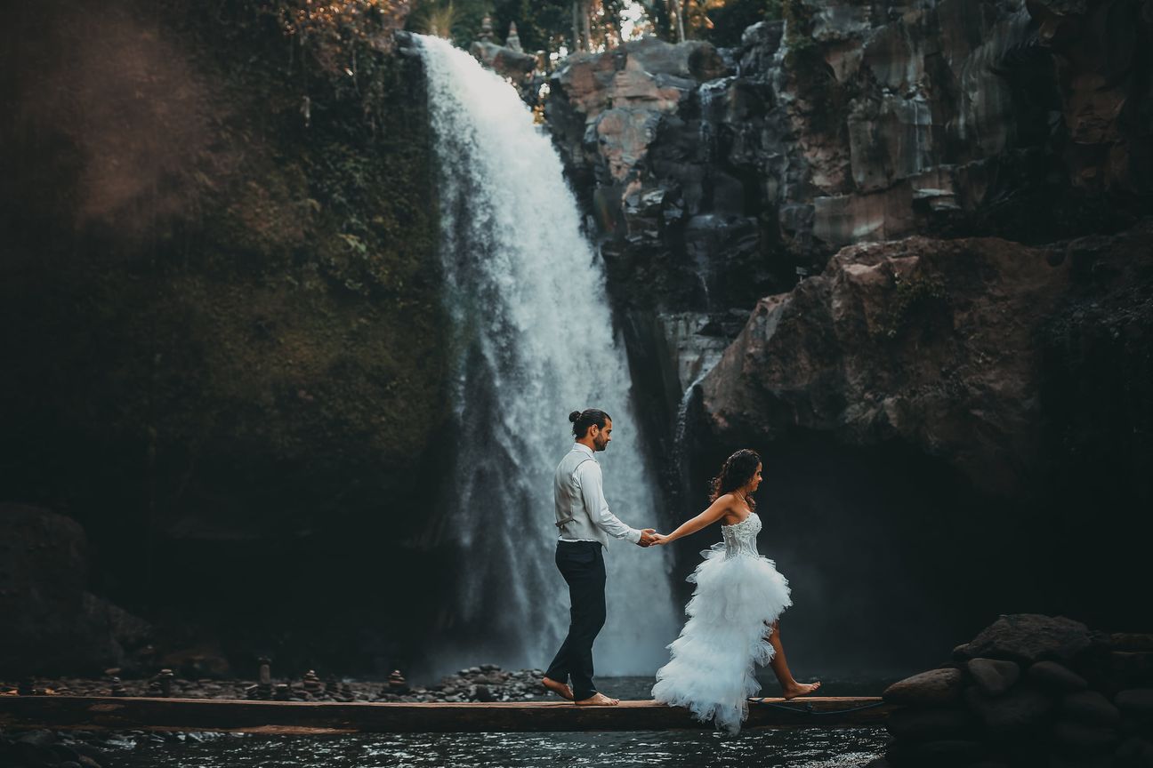 A newlywed couple pose together beneath a tall forest waterfall