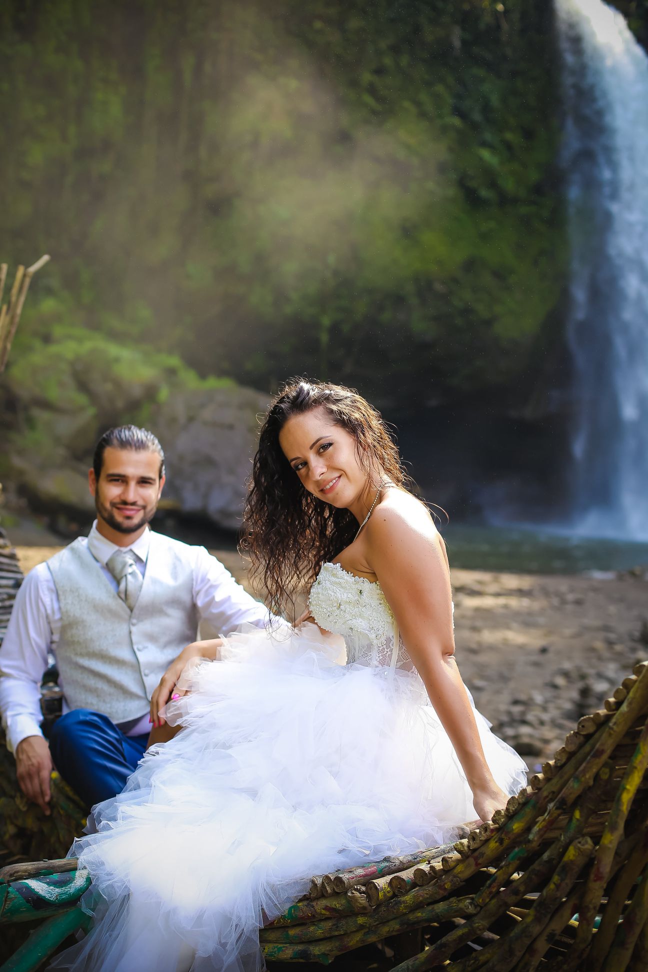 A newlywed couple sit together beside a forest waterfall