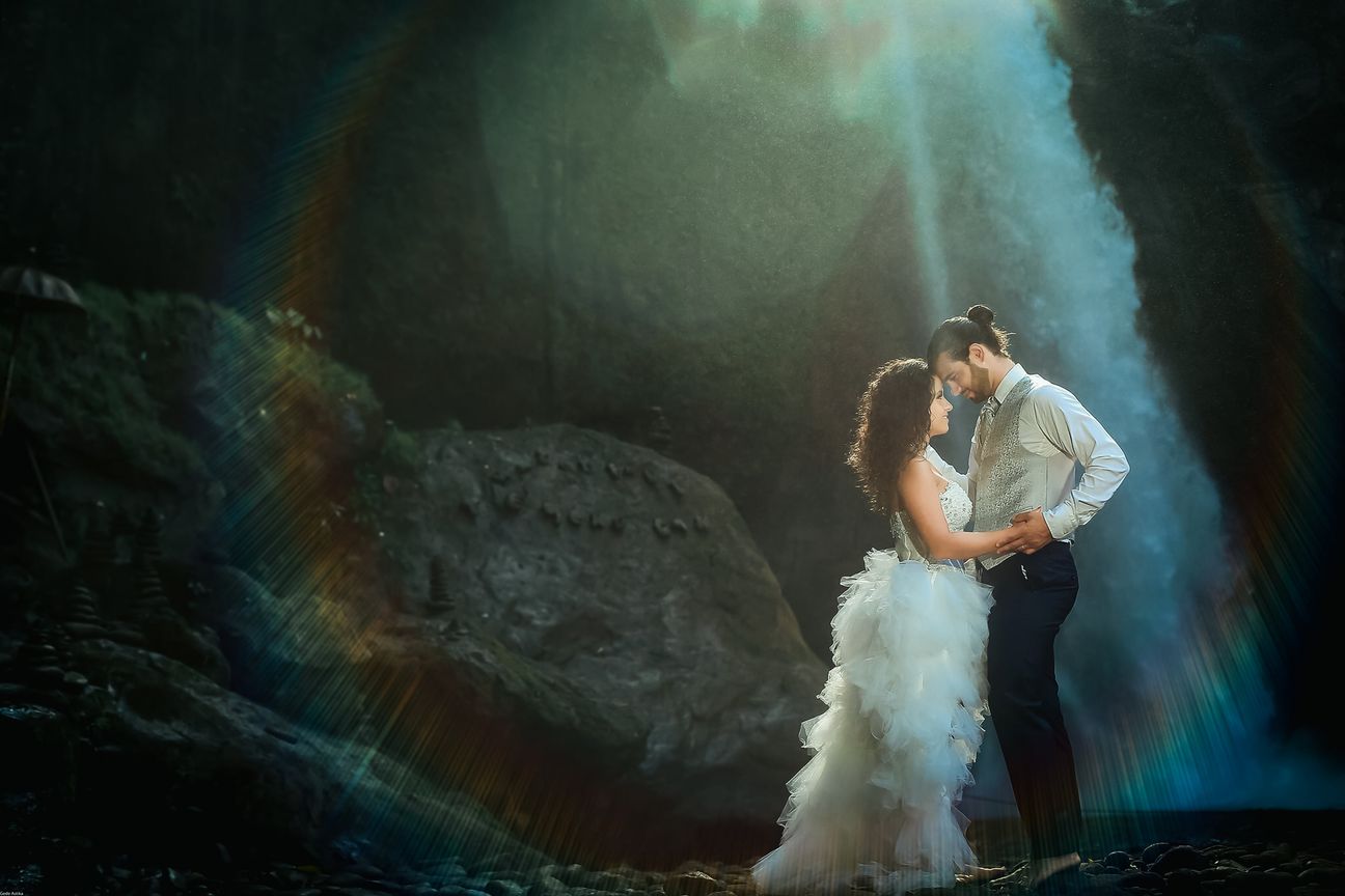 A newlywed couple pose together beneath a tall forest waterfall