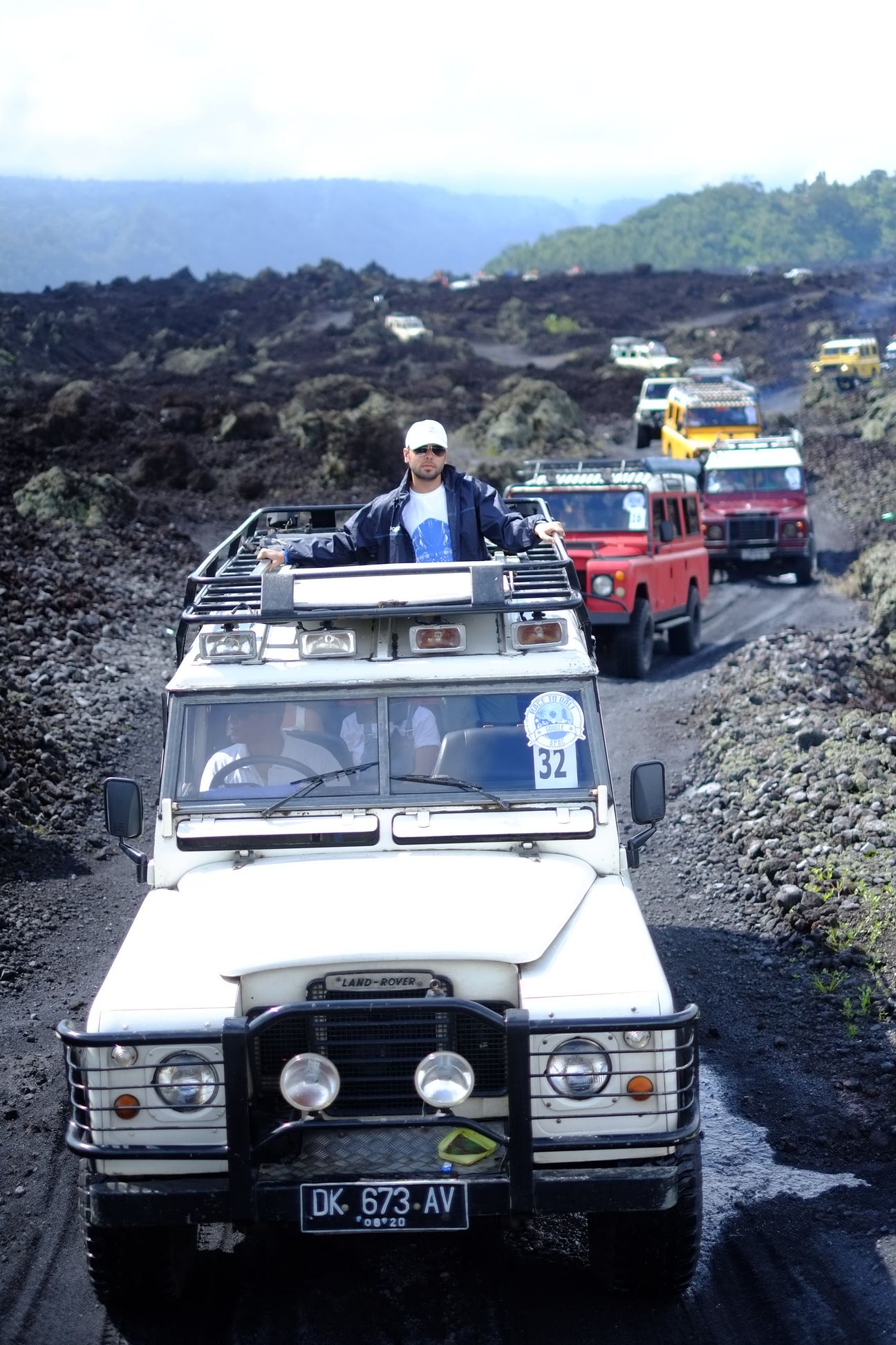 A man stands through the roof of a white Land Rover leading a line of jeeps along a black lava track