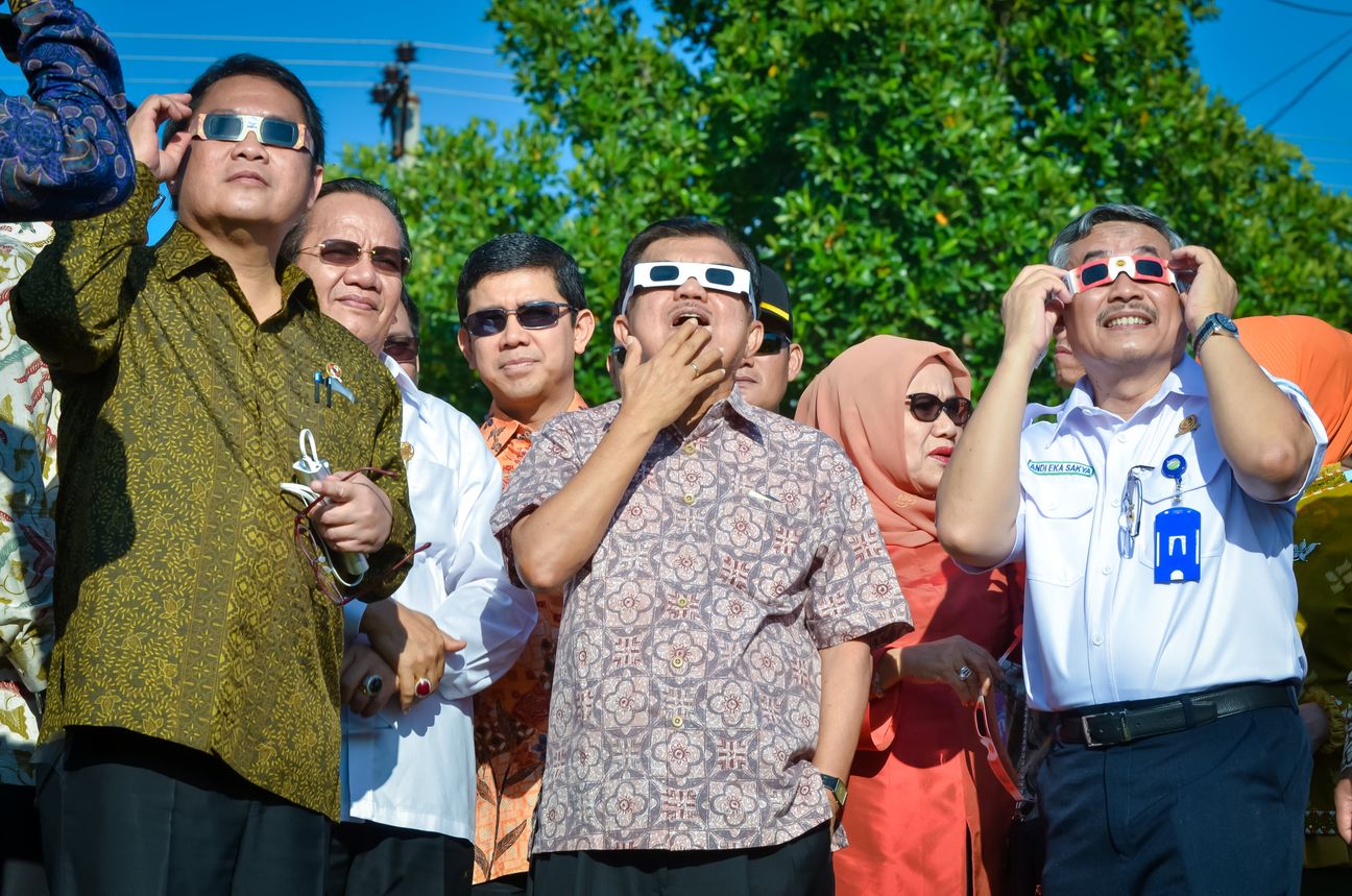 A row of guests in batik tip their heads back behind cardboard eclipse glasses