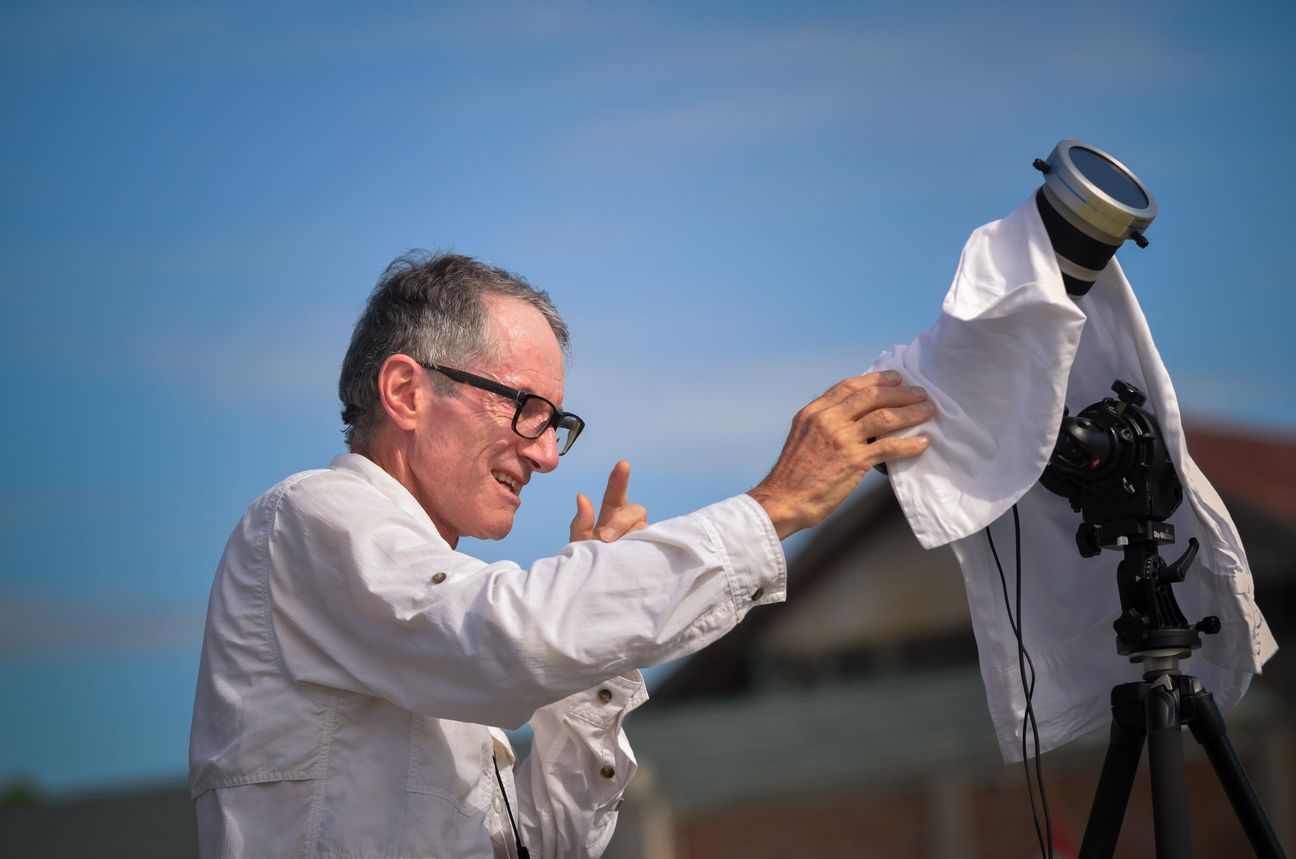 A man pulls a white shirt over a telescope and camera on a tripod against a blue sky