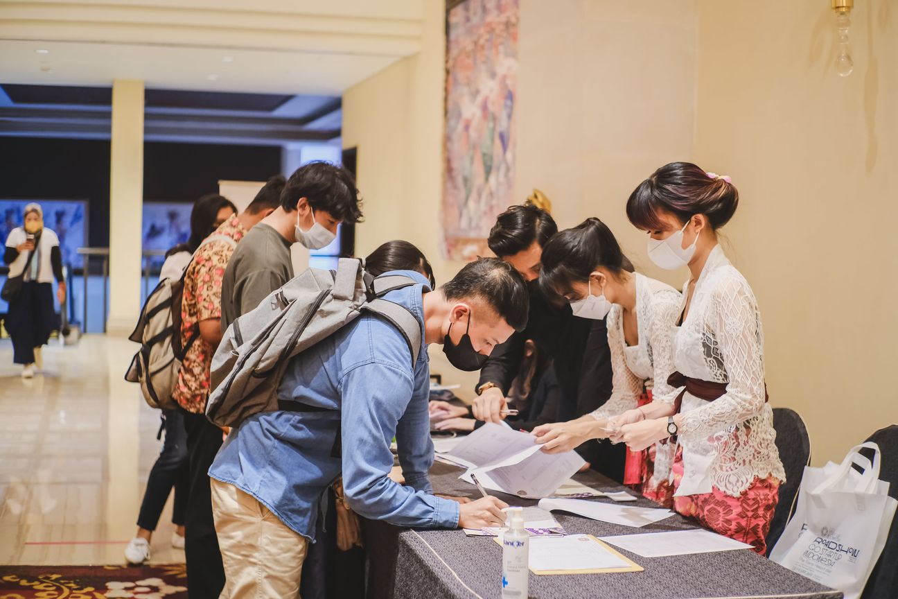 Masked delegates bend over the registration desk while hosts in kebaya hand out papers
