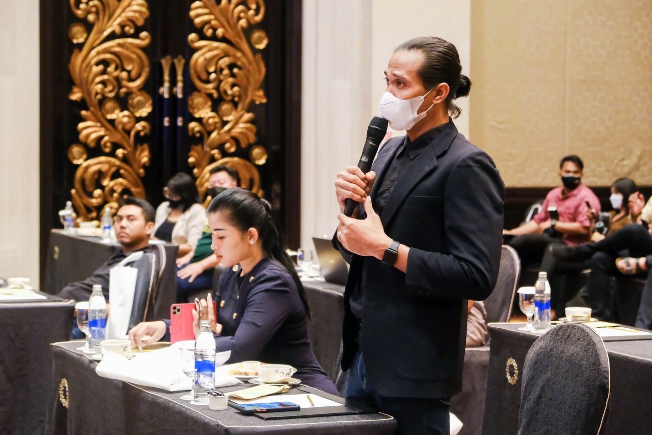 A delegate in a black jacket stands to speak, tables of listeners around him