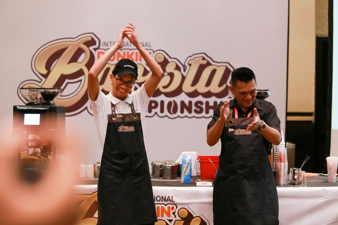 A barista throws both arms up beside the espresso bar as another applauds