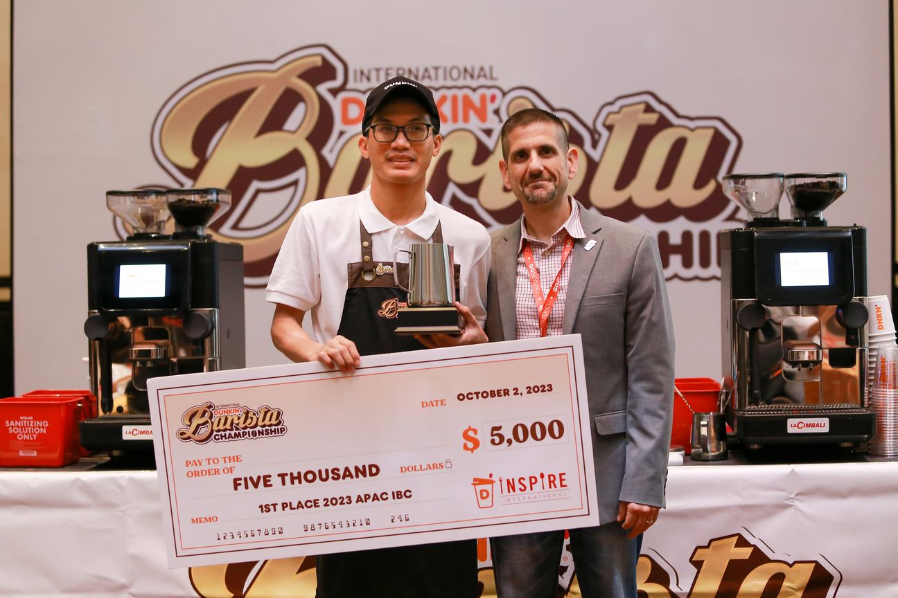 A barista holds a trophy pitcher and an oversized cheque beside an organiser