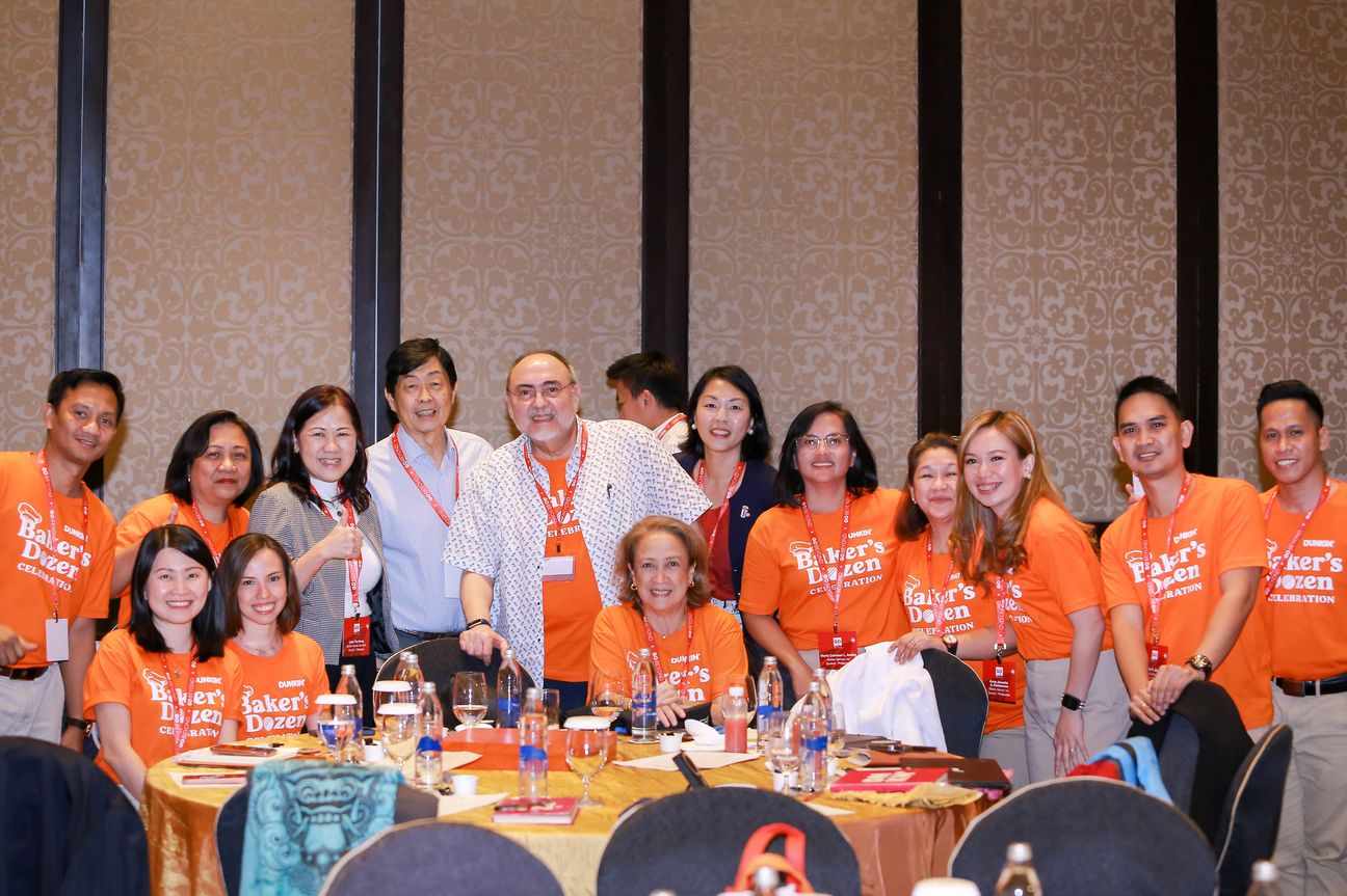 A team in orange celebration shirts crowd in around two guests at their table