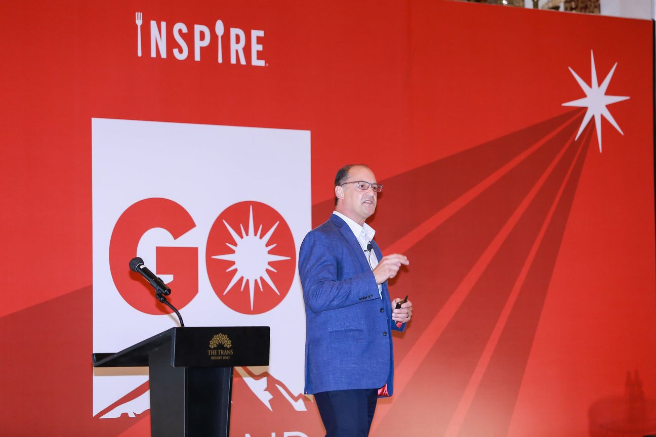 A speaker in a blue jacket presents from a lectern against a red backdrop