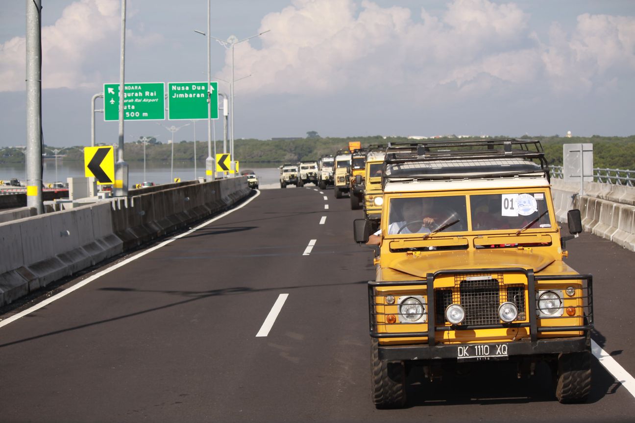 A yellow Land Rover leads the convoy along the bypass under a high afternoon sky