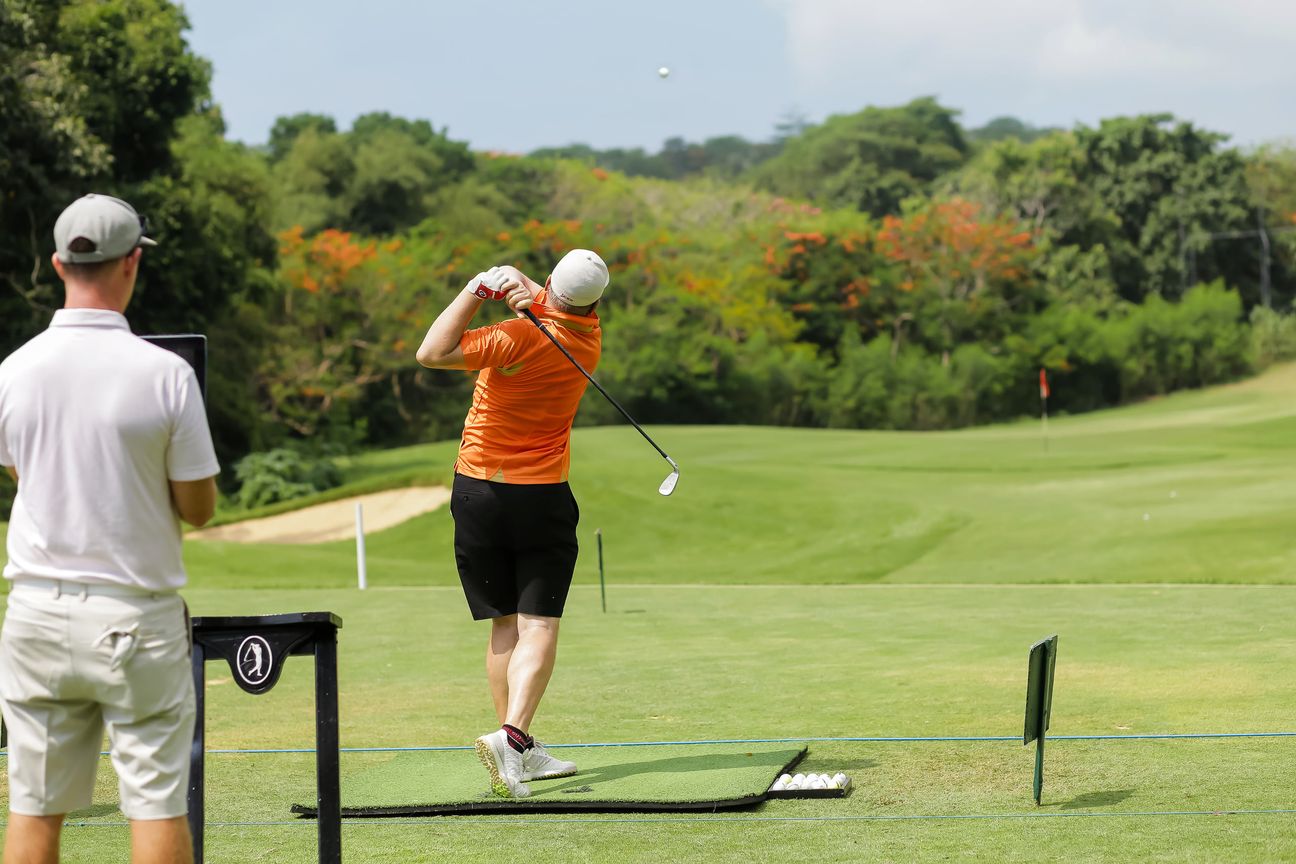 A golfer in orange swings on the practice range while a coach watches