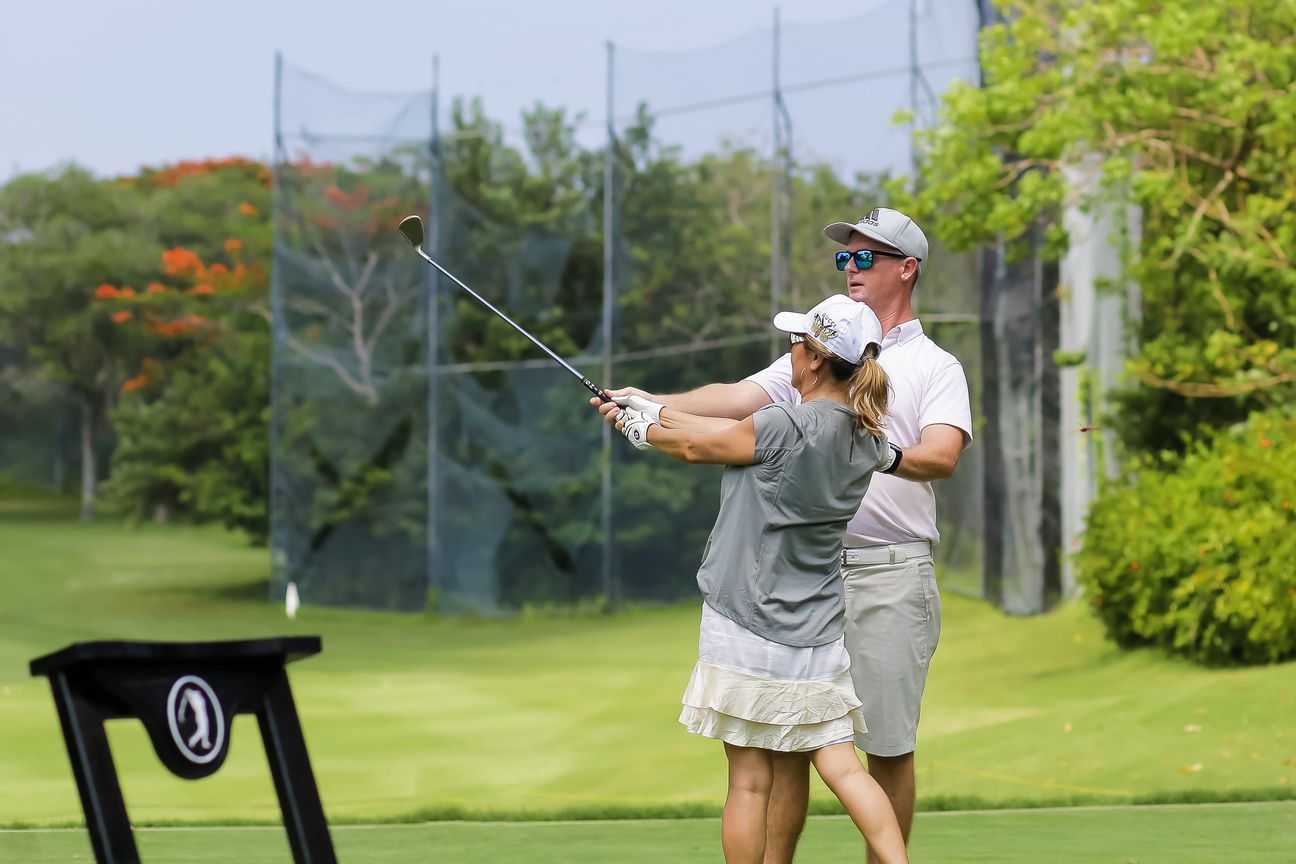 A coach stands close behind a golfer as she holds the finish of her swing
