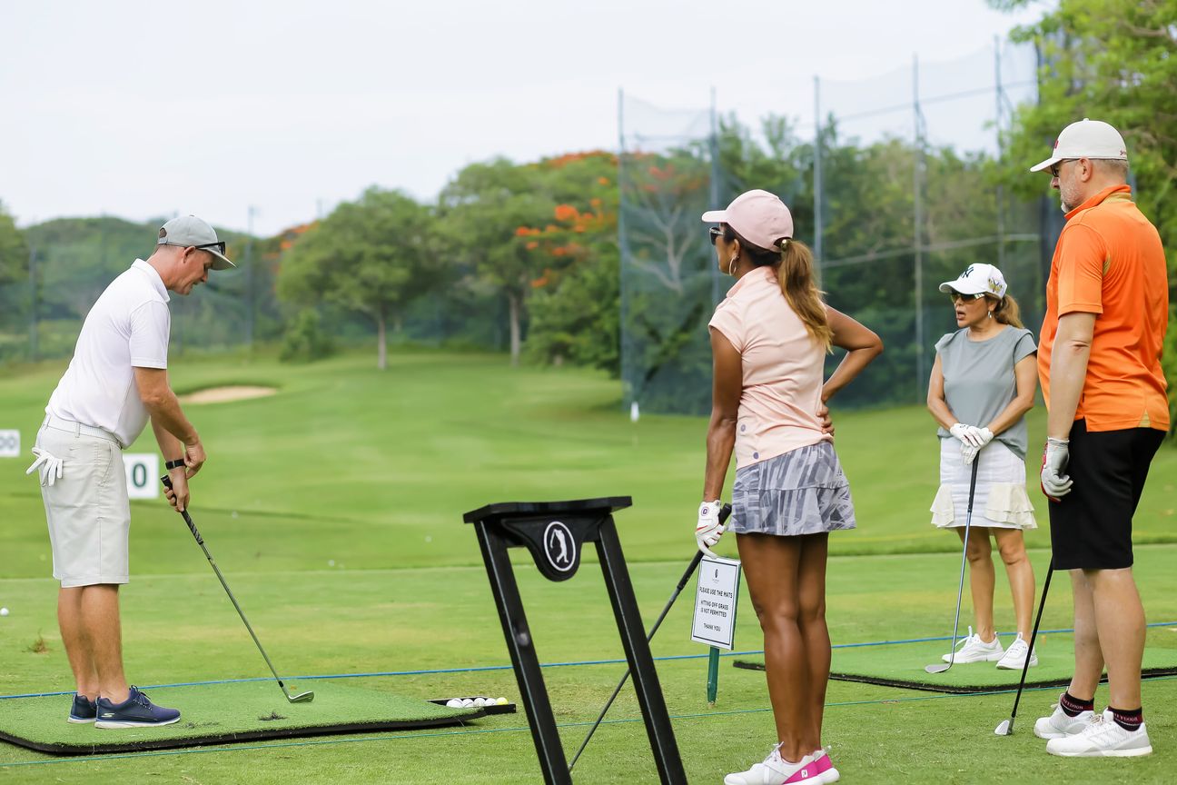 Three golfers watch a fourth play across the practice mats