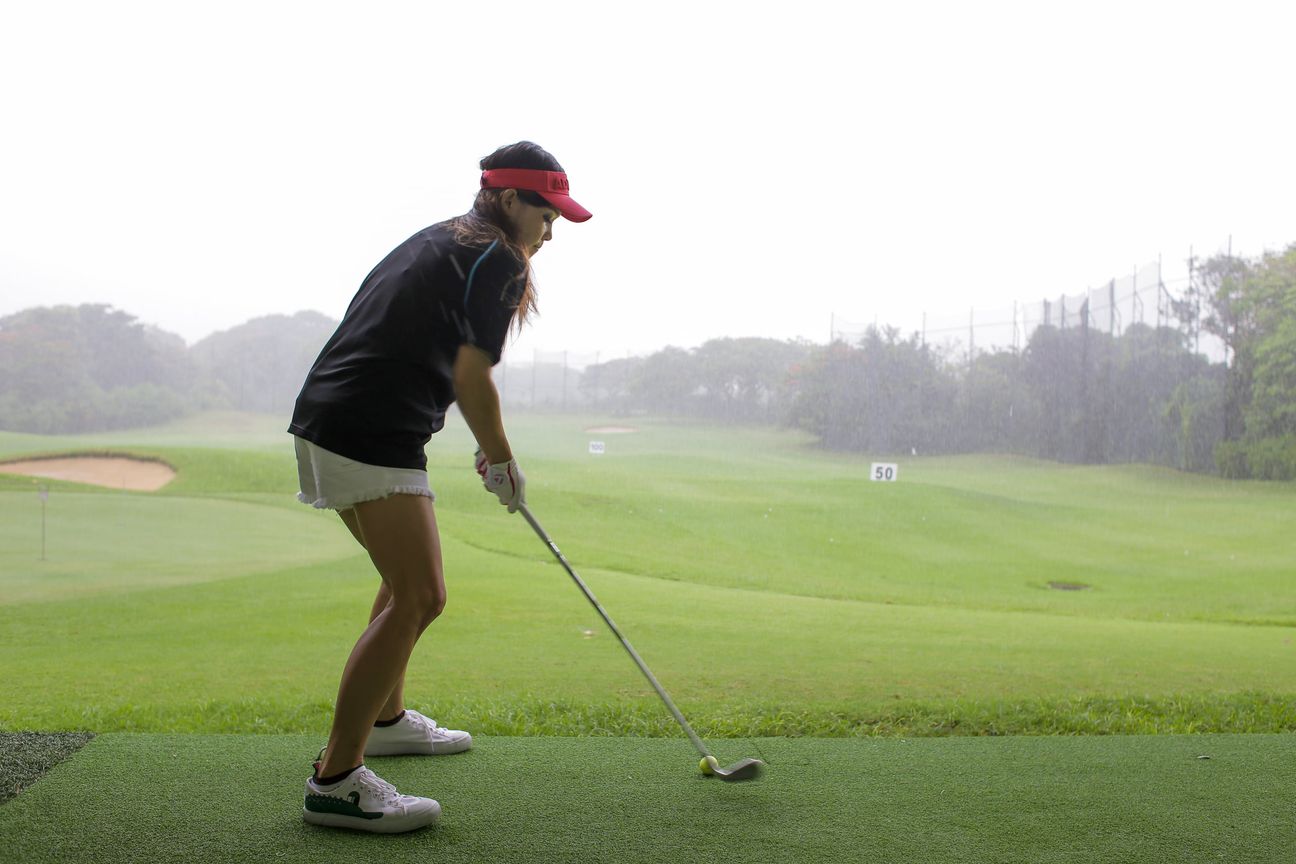 A golfer lines up a drive on a wet range as rain streaks across the frame