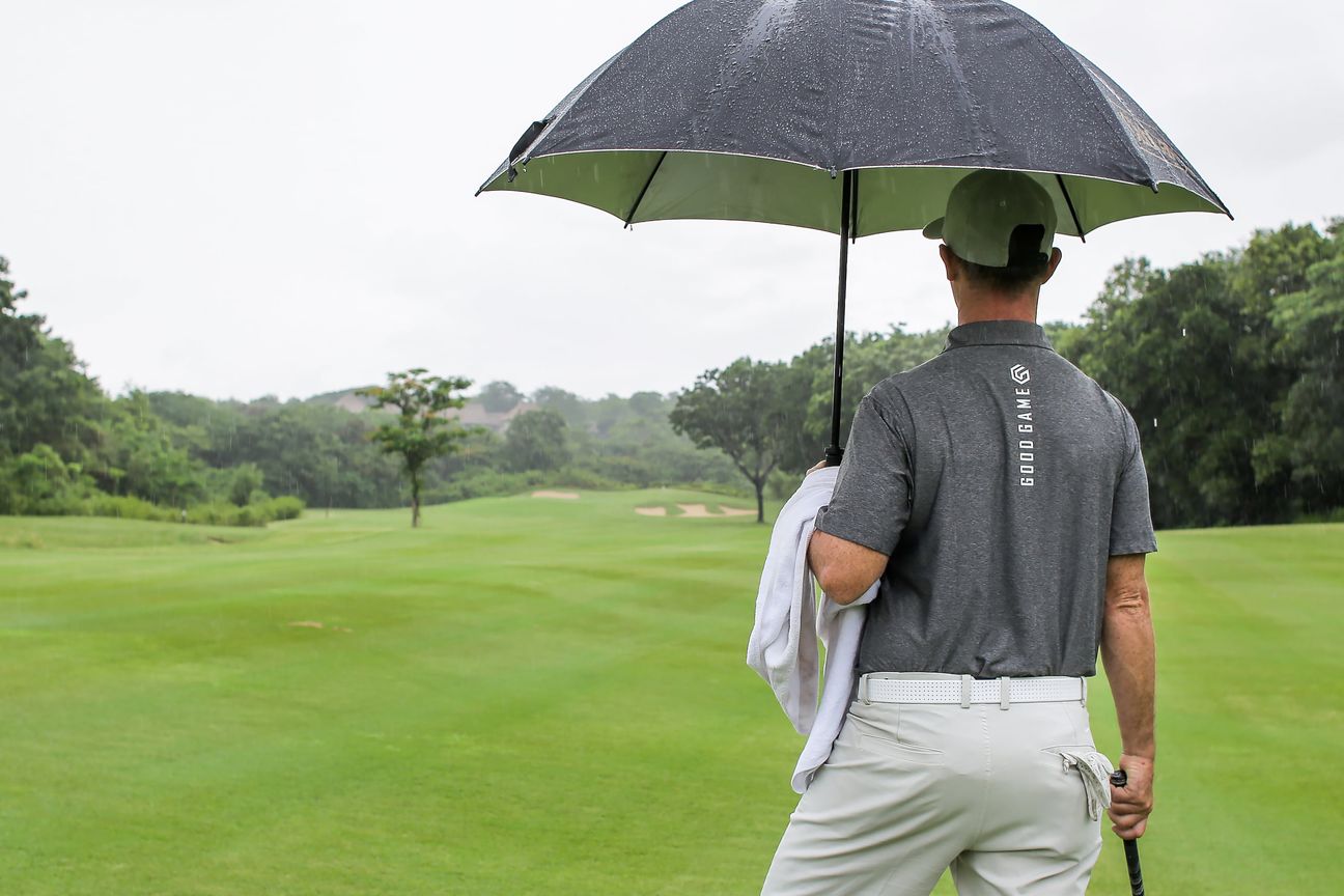 A golfer stands under an umbrella looking down an empty fairway in the rain