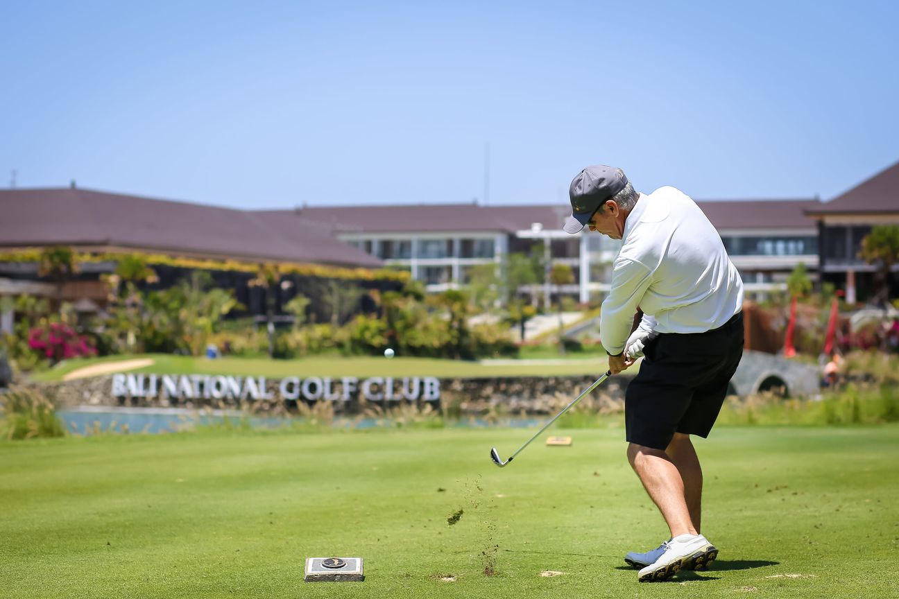 A golfer strikes the ball and lifts a divot in front of the clubhouse