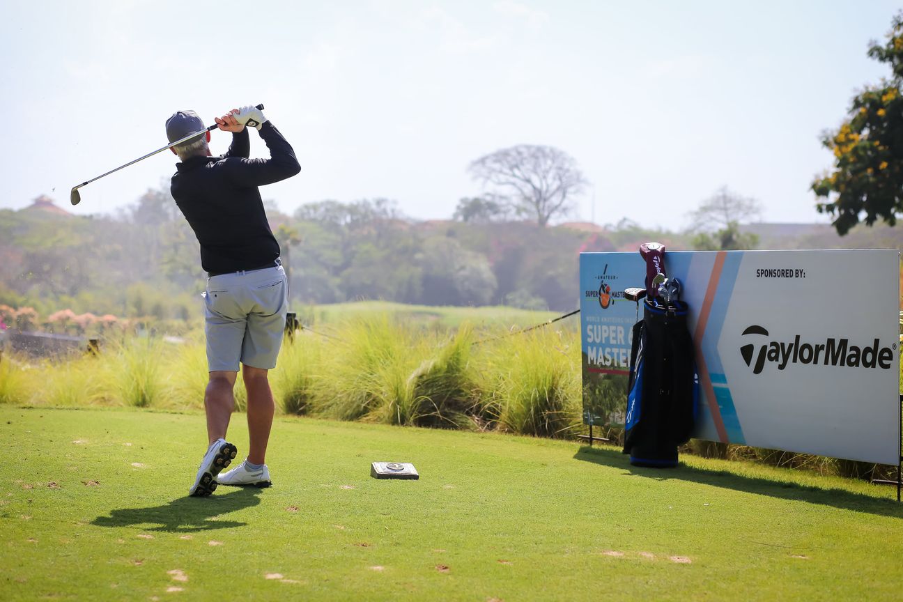 A golfer holds the finish of a drive, his bag standing beside the tee