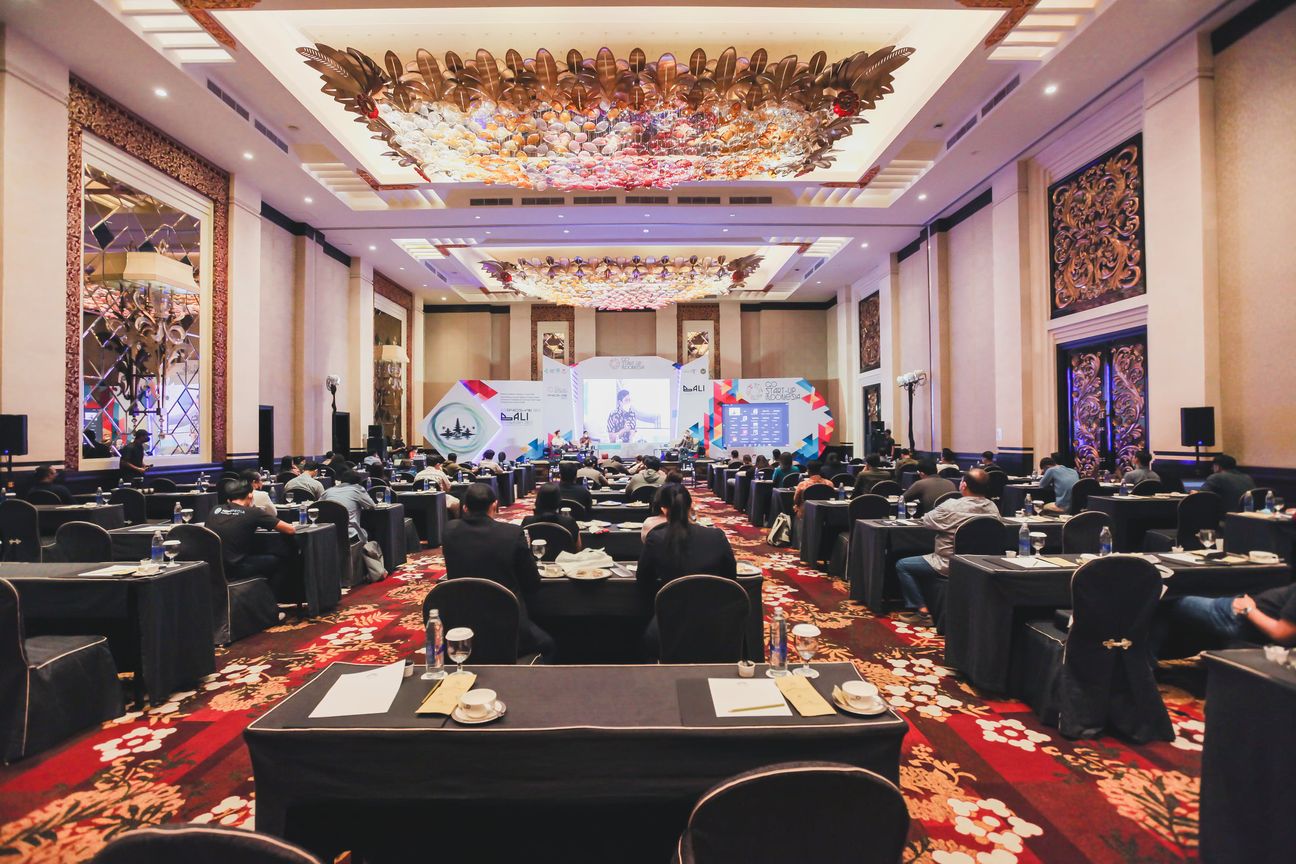 Rows of draped tables in a Balinese ballroom, delegates watching the screen