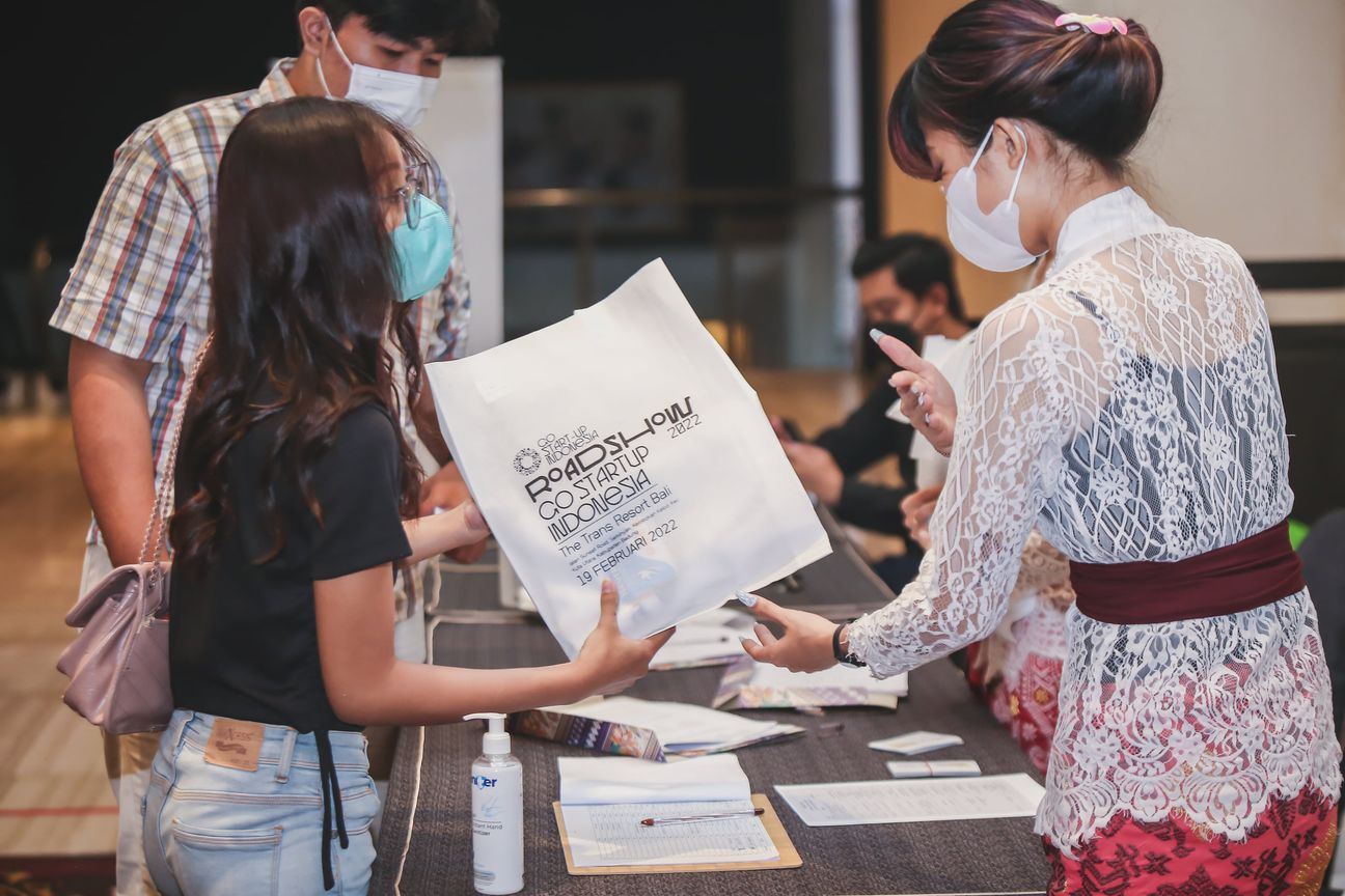 A host in lace kebaya passes a printed tote bag across the registration desk