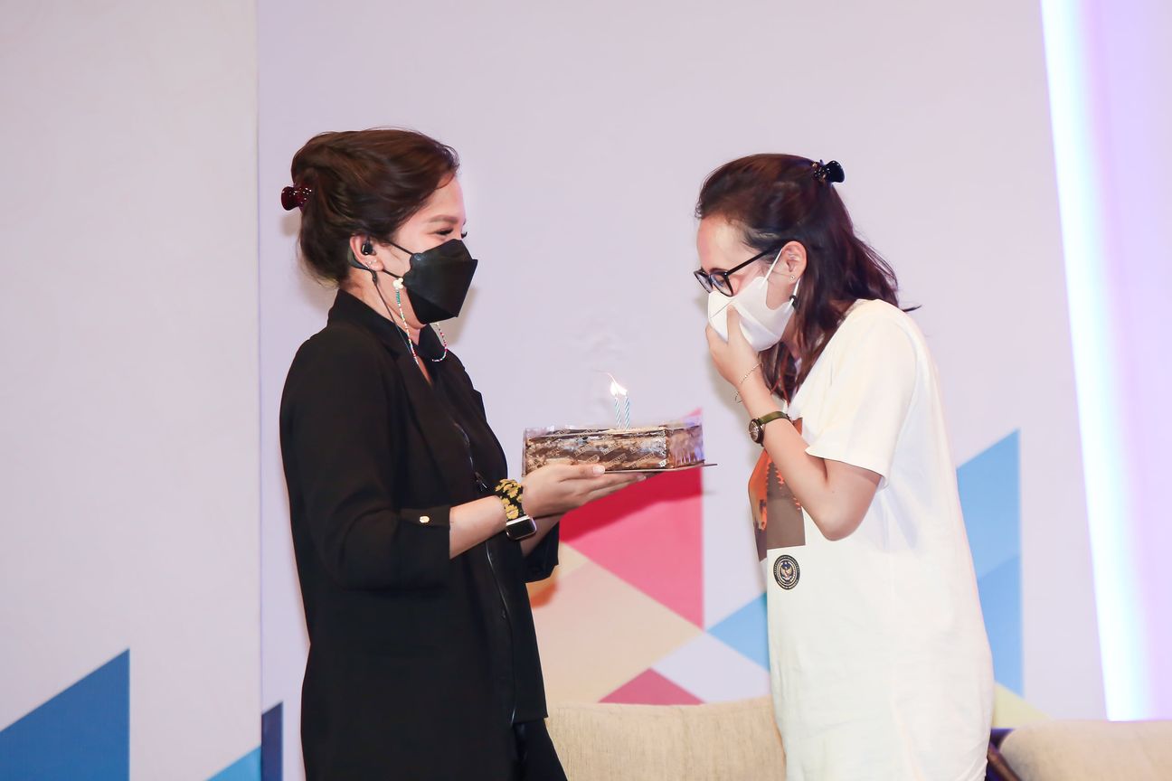 One woman holds out a small cake with a lit candle to another, who covers her face