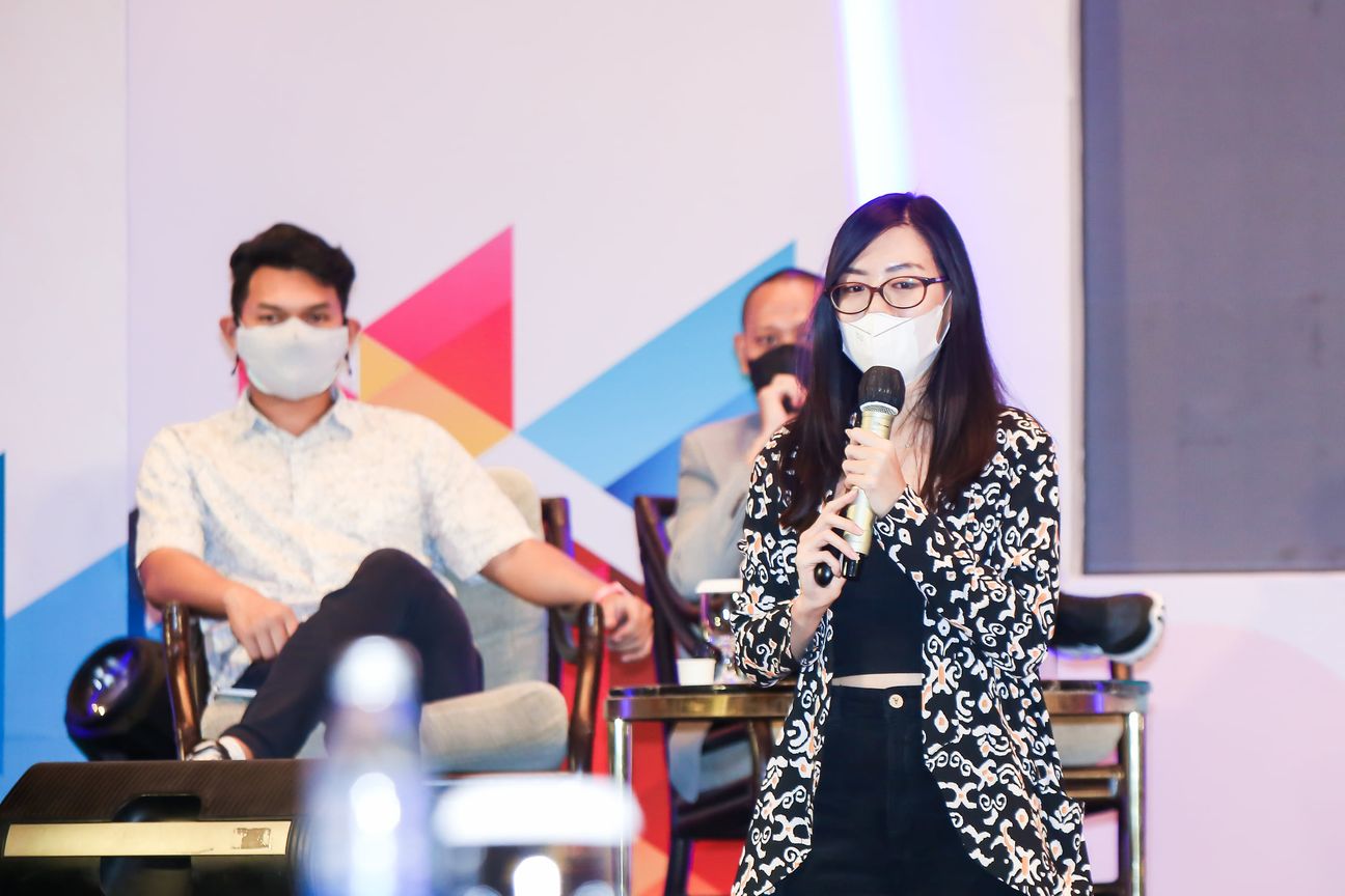 A panellist in batik speaks into a microphone on stage while another listens beside her