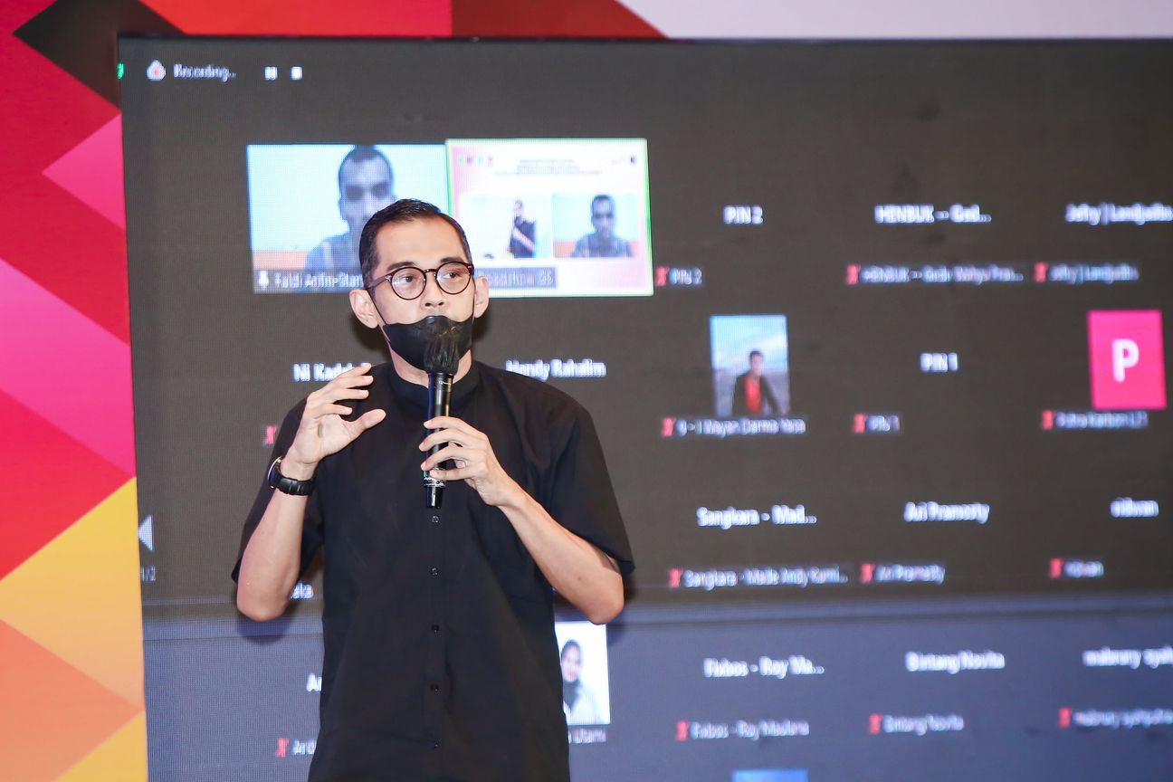 A speaker in black addresses the hall in front of a projected grid of remote attendees