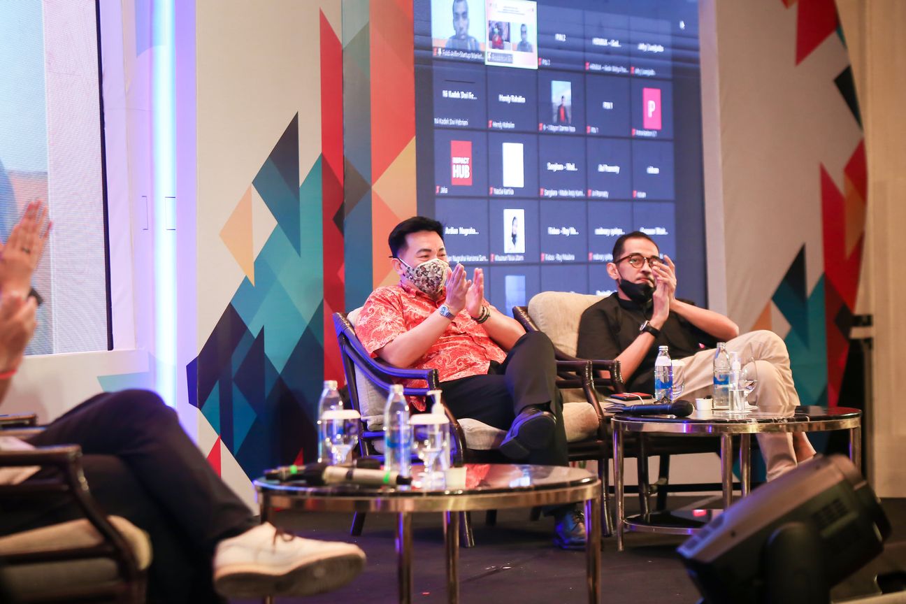 Two panellists applaud from armchairs on stage, the video call still projected behind them