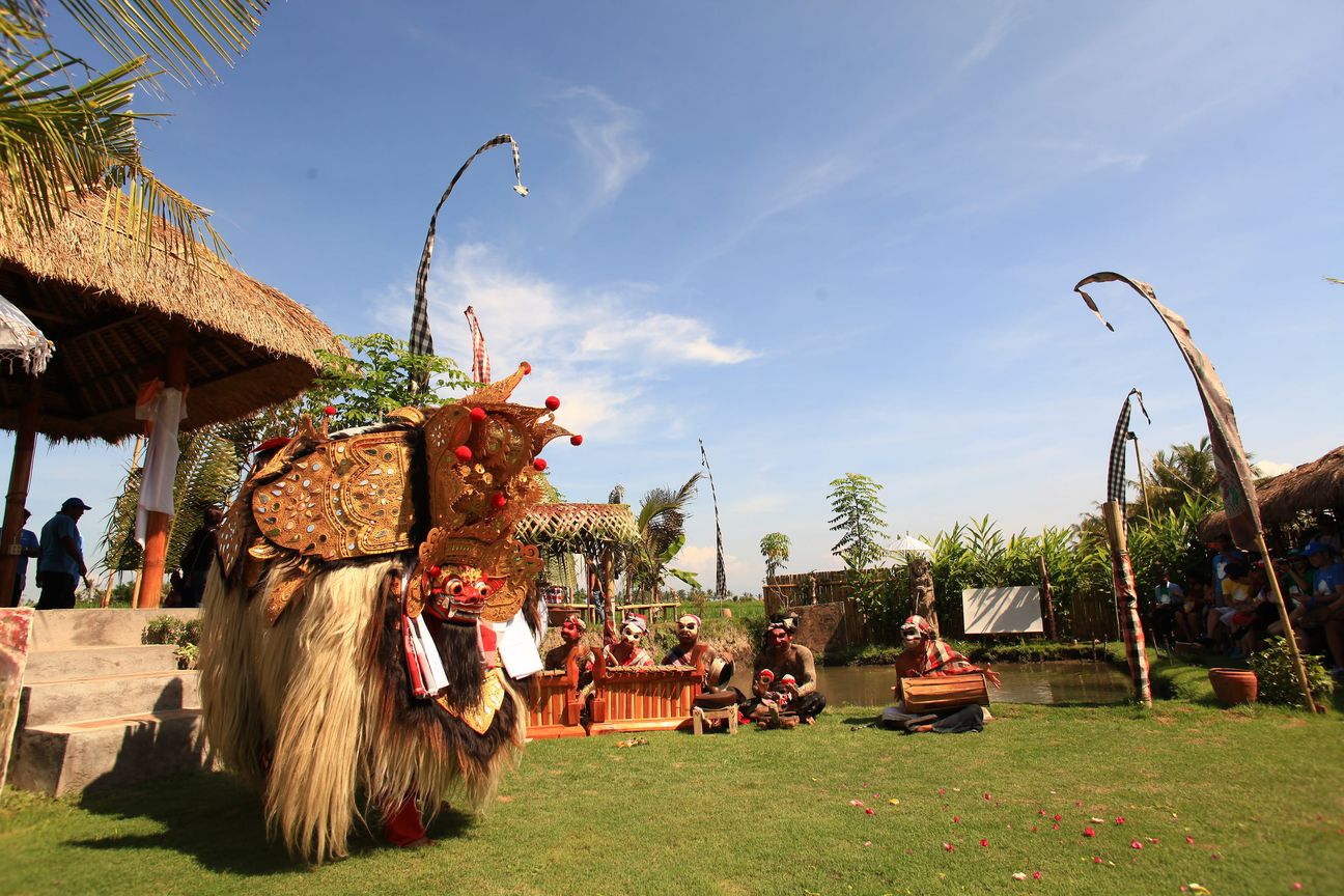 A Barong costume waits on a lawn beside a gamelan ensemble, penjor poles bending overhead