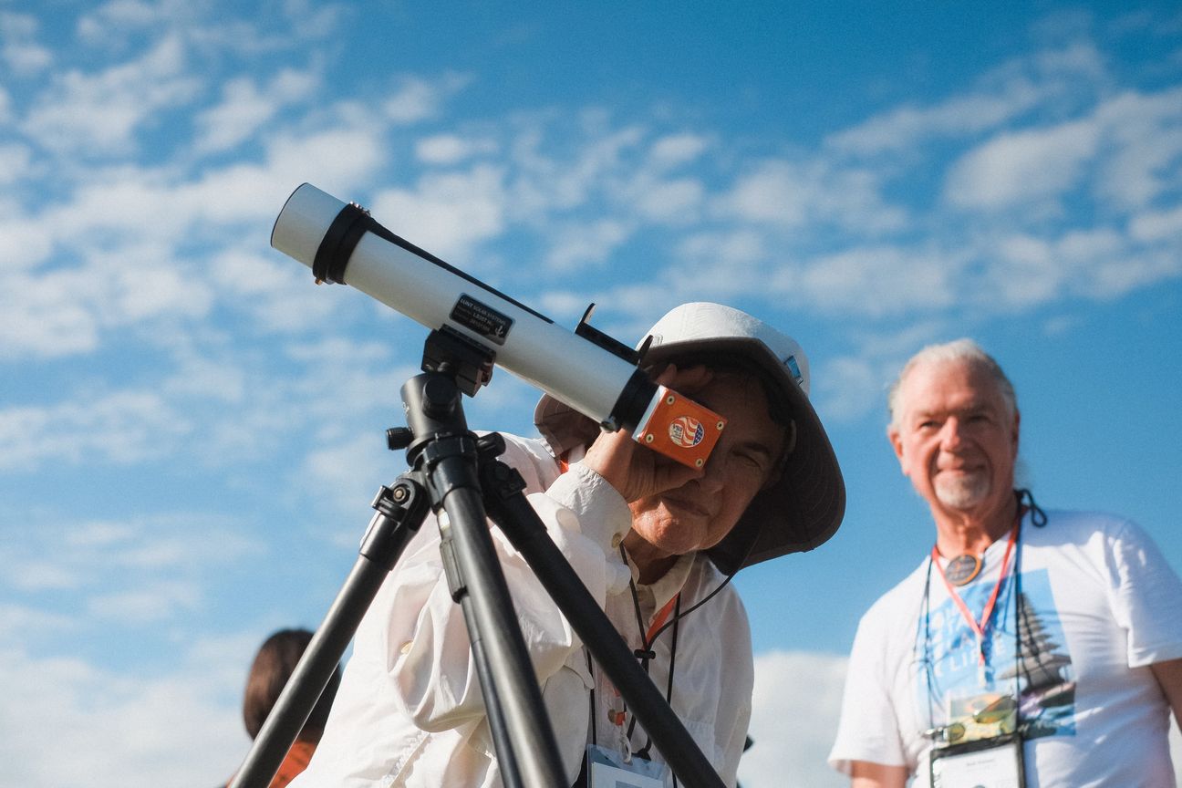 A woman in a sun hat squints into a small telescope while a man beside her watches