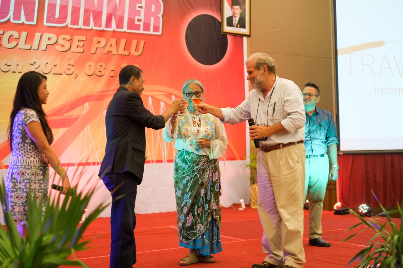 Three guests reach in to touch glasses on stage in front of an eclipse banner